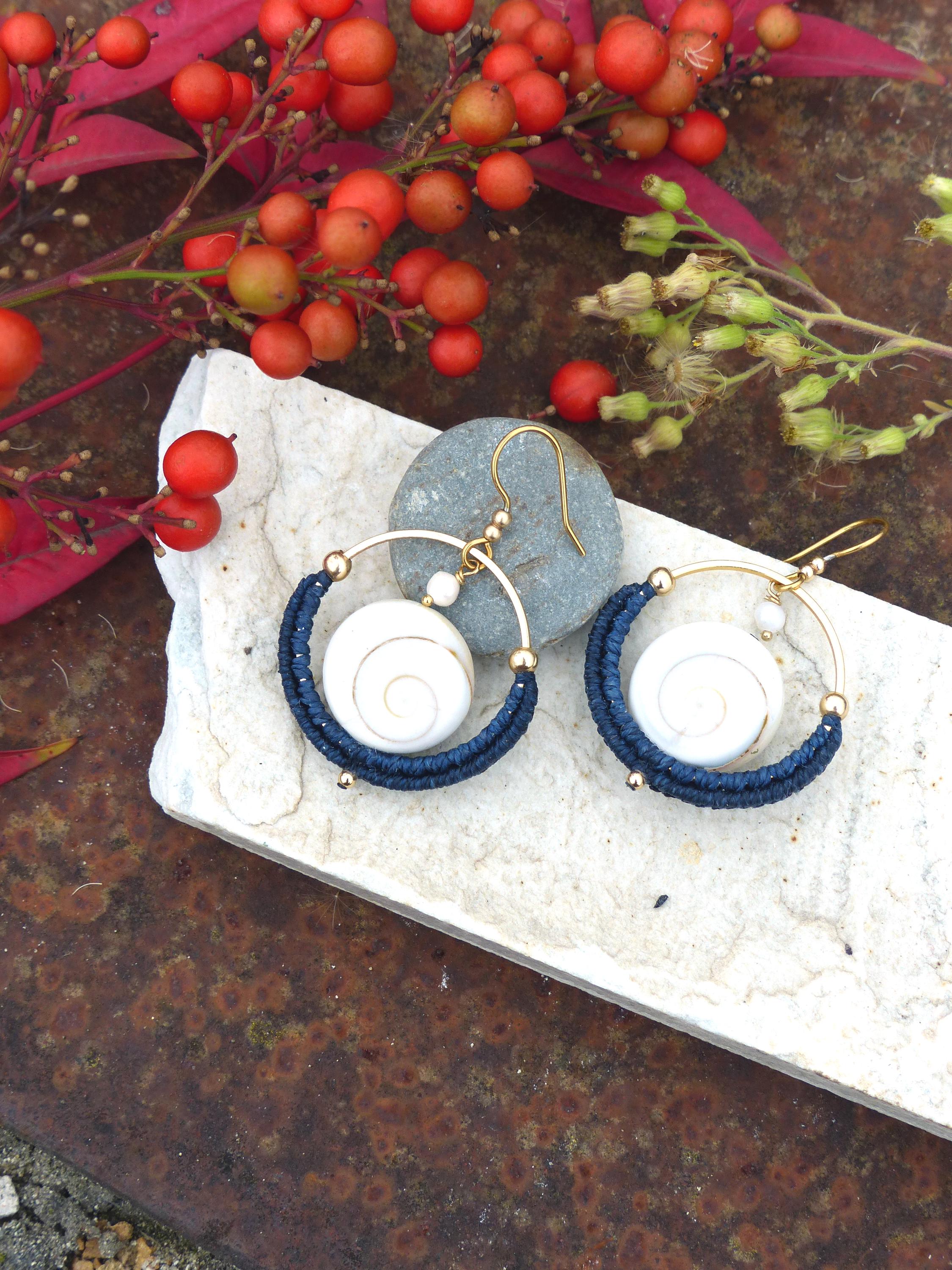 Peut inclure: Boucles d'oreilles faites à la main avec des cerceaux dorés, des spirales blanches et des détails en fil bleu marine. Les boucles d'oreilles sont présentées sur une surface en pierre blanche, avec des baies rouges et du feuillage en arrière-plan.