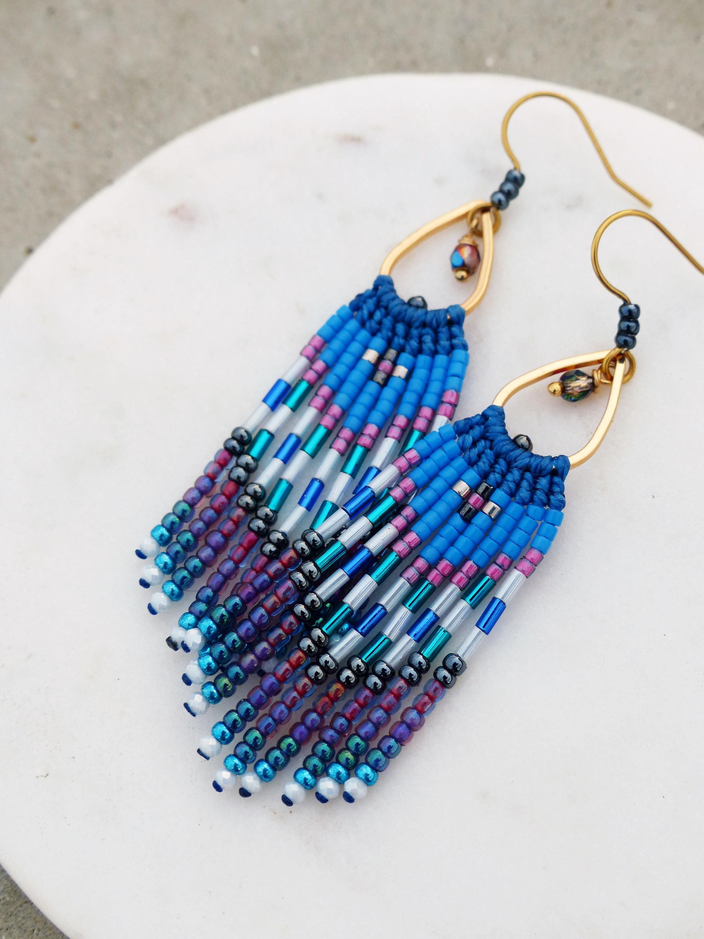 Peut inclure: Une paire de boucles d'oreilles faites à la main avec un motif de franges. Les boucles d'oreilles présentent un cadre en forme de goutte d'eau de couleur dorée et sont ornées de perles bleues, violettes, argentées et blanches. La frange est faite de plusieurs brins de perles.