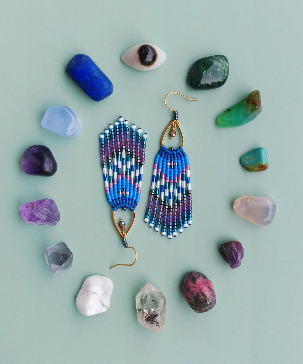 Peut inclure: Boucles d'oreilles bleues en perles avec des crochets et des accents dorés, entourées de diverses pierres précieuses et cristaux colorés. Les boucles d'oreilles présentent un motif géométrique avec des perles blanches, bleues et violettes. Le fond est vert clair.