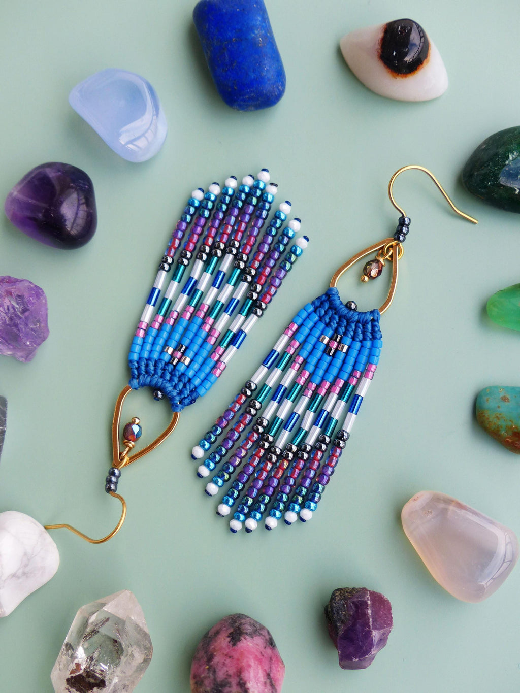 Peut inclure: Boucles d'oreilles faites à la main avec des perles bleues, blanches, roses et violettes. Les boucles d'oreilles ont un crochet doré et un motif en forme de goutte. Diverses pierres précieuses colorées entourent les boucles d'oreilles sur une surface vert clair.