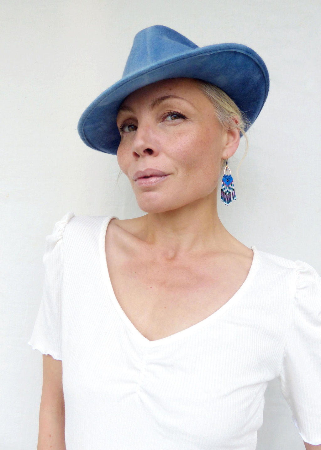 Peut inclure: Une femme porte un chapeau bleu et un haut blanc à col en V. Elle porte également des boucles d'oreilles en perles avec des accents bleus, blancs et violets. Le chapeau a un large bord et une texture douce.