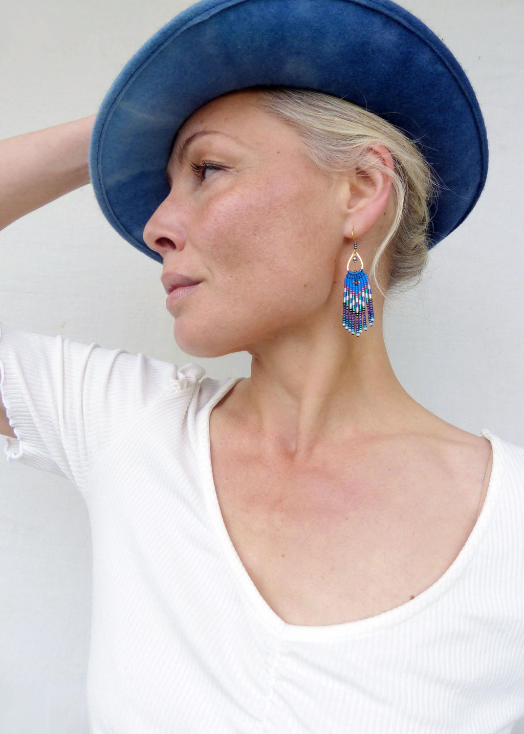 Peut inclure: Une femme porte un chapeau bleu et un haut blanc. L'image met l'accent sur des boucles d'oreilles en perles avec un dessus doré et une frange bleue, blanche et violette.