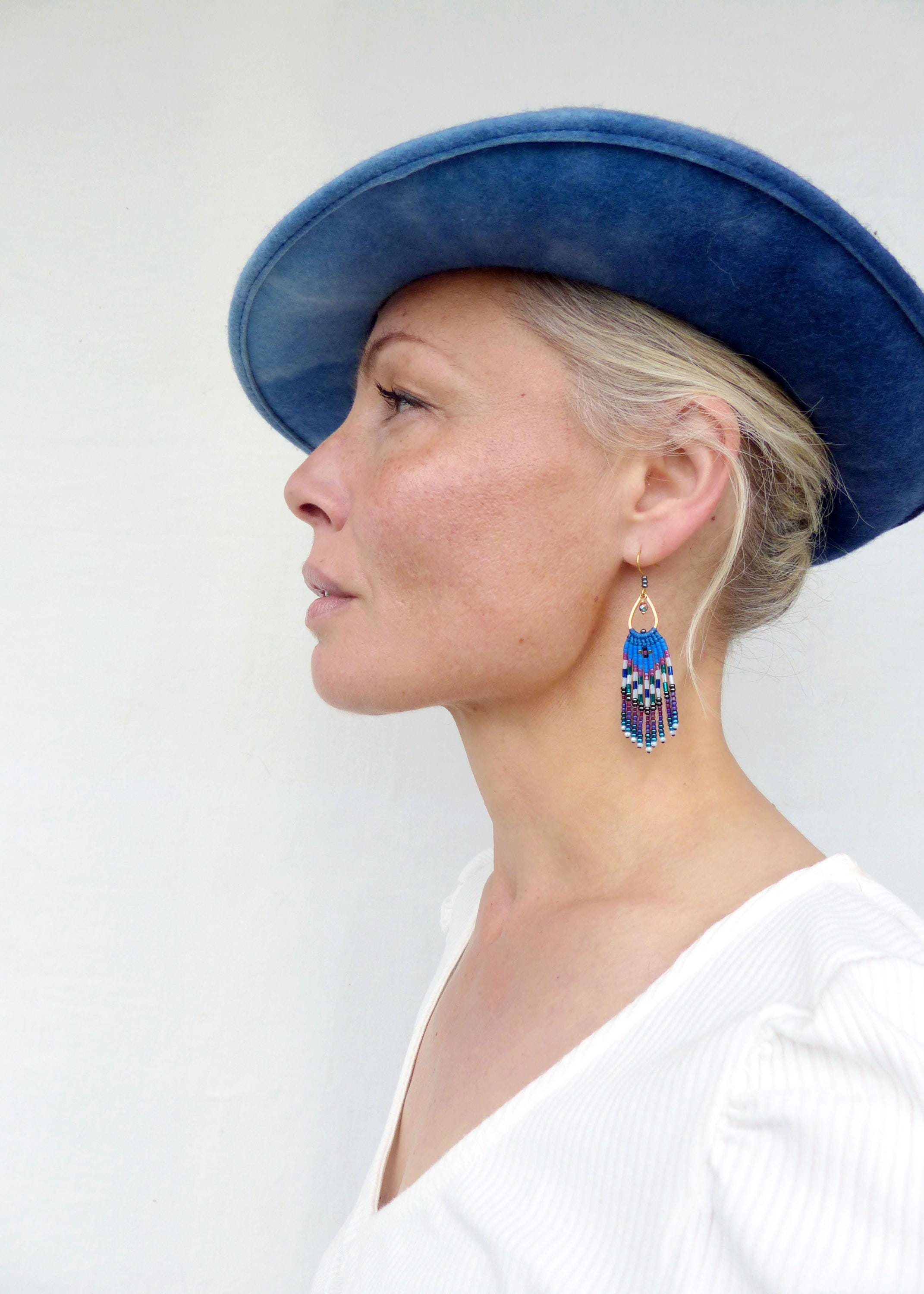 Peut inclure: Profil d'une personne portant un chapeau bleu et une chemise blanche, avec des boucles d'oreilles en perles. Les boucles d'oreilles ont un dessus doré et présentent un motif de perles bleues, violettes et blanches, créant un effet de frange. Le fond est blanc.