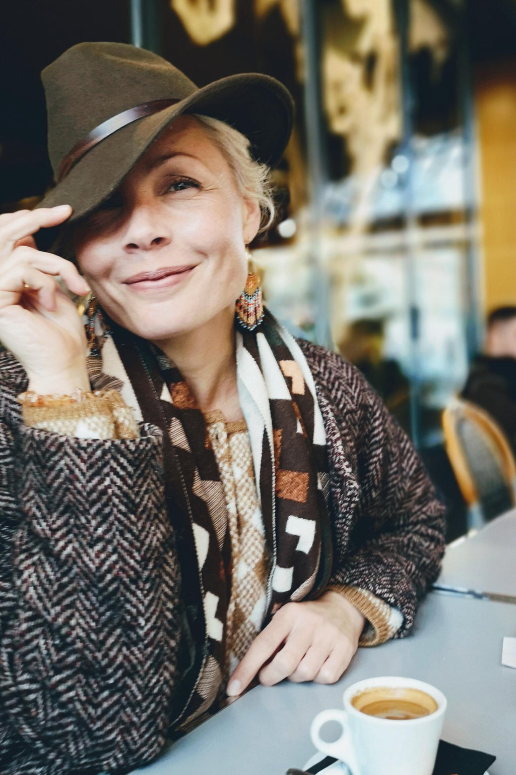 Peut inclure: Une femme porte un chapeau fedora marron avec une bande en cuir, une écharpe à motifs et un manteau en tweed marron. Elle sourit et tient le bord du chapeau. Une tasse de café blanc est sur la table.
