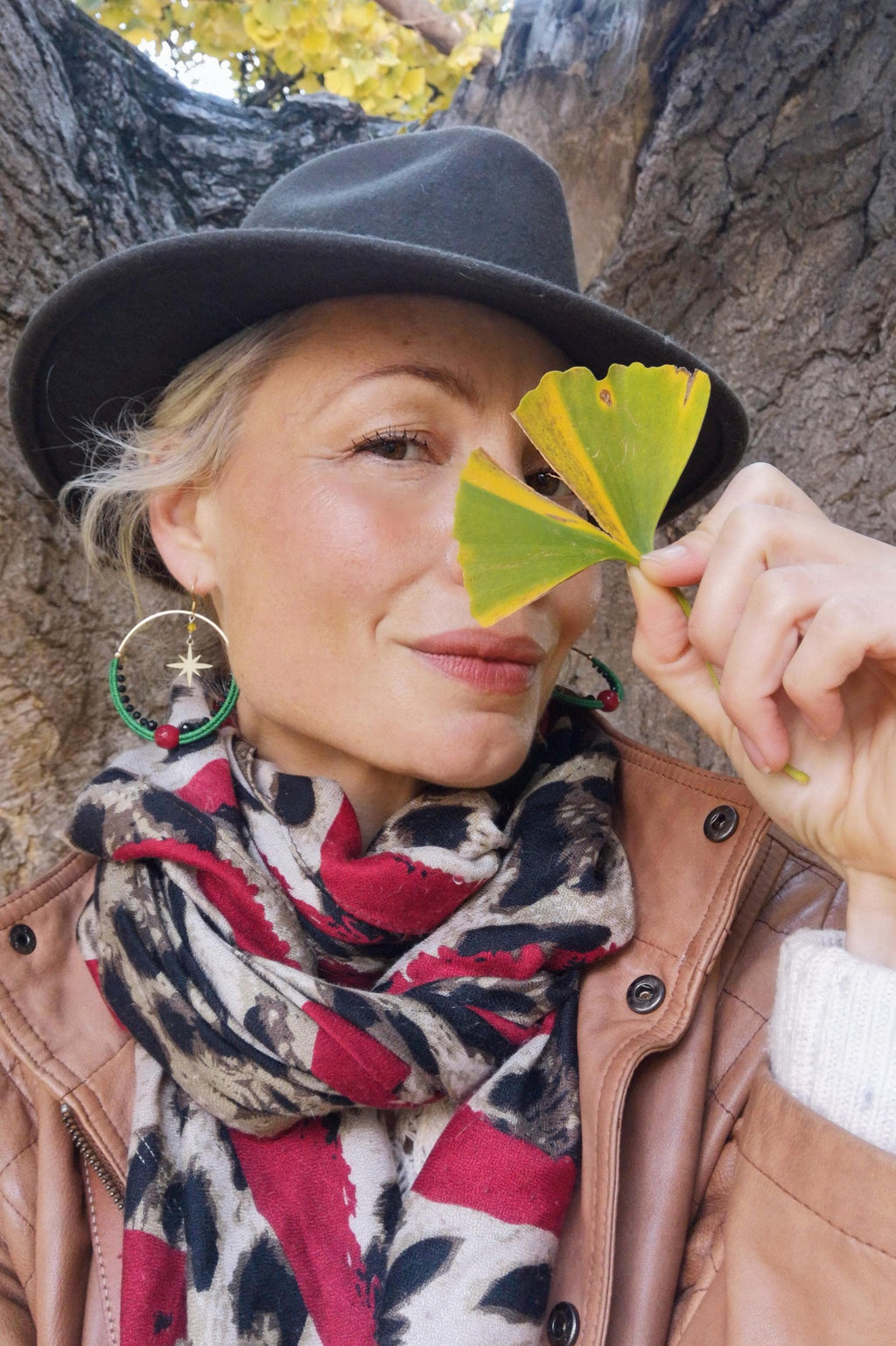 Peut inclure: Une femme porte un chapeau fedora gris foncé, des boucles d'oreilles créoles dorées avec un charme en forme d'étoile et une écharpe à motifs rouges et noirs. Elle tient une feuille jaune et verte devant son visage. Elle porte une veste en cuir marron.