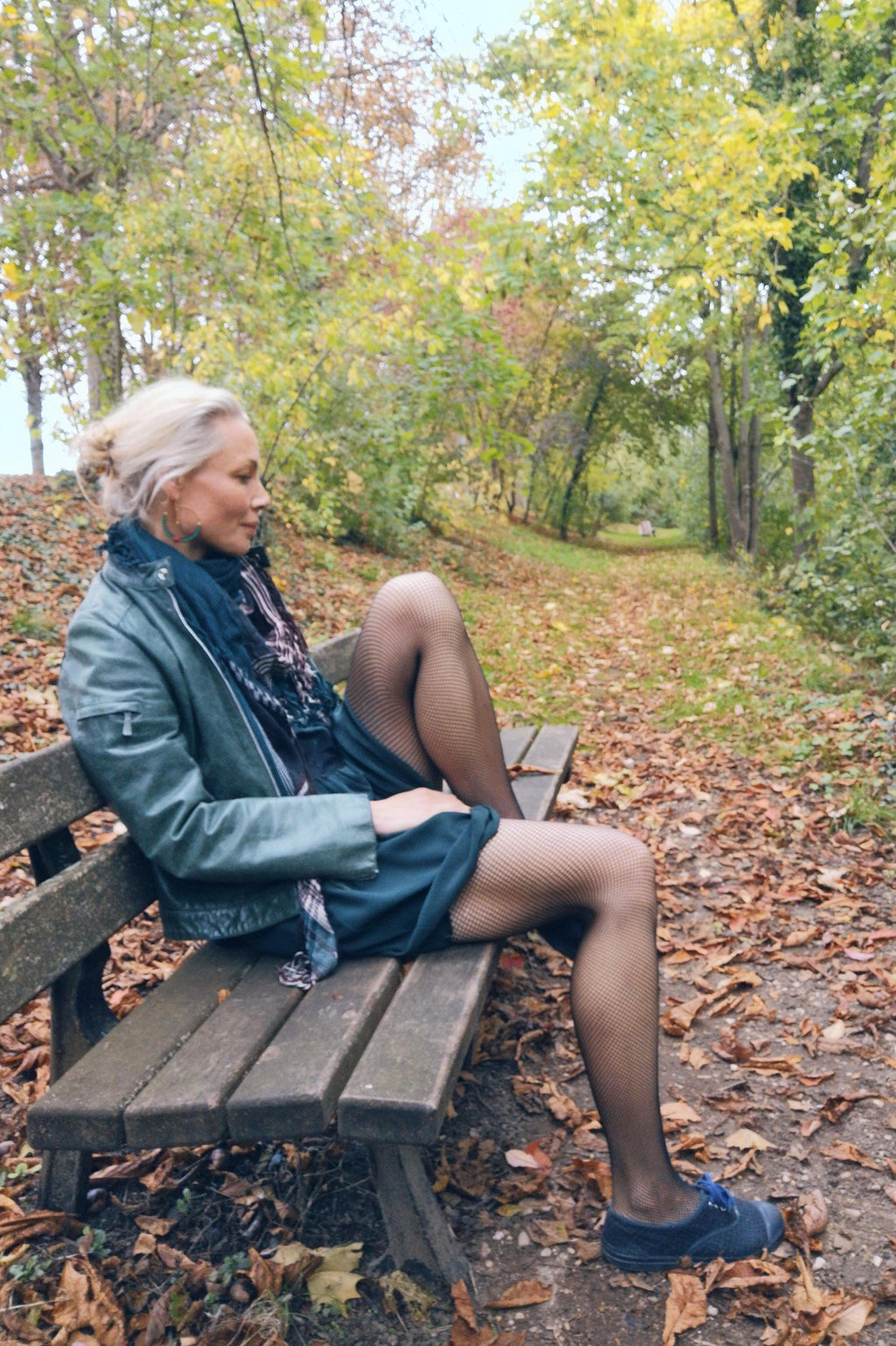 Peut inclure: Une personne assise sur un banc en bois dans un parc. Elle porte une veste en cuir vert d'eau, une écharpe bleu foncé et une robe vert d'eau. Des bas résille noirs et des chaussures bleues complètent la tenue. Le sol est couvert de feuilles mortes.
