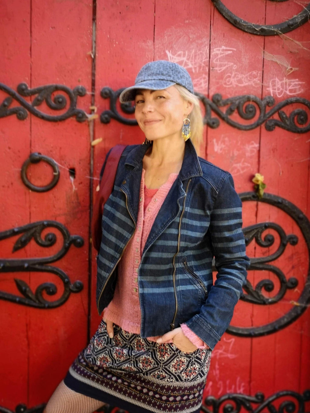Peut inclure: Veste en jean à rayures horizontales bleues, portée sur un pull rose et une jupe à motifs. La personne porte une casquette de baseball en jean et des boucles d'oreilles. L'arrière-plan est une porte rouge avec des ferronneries noires.