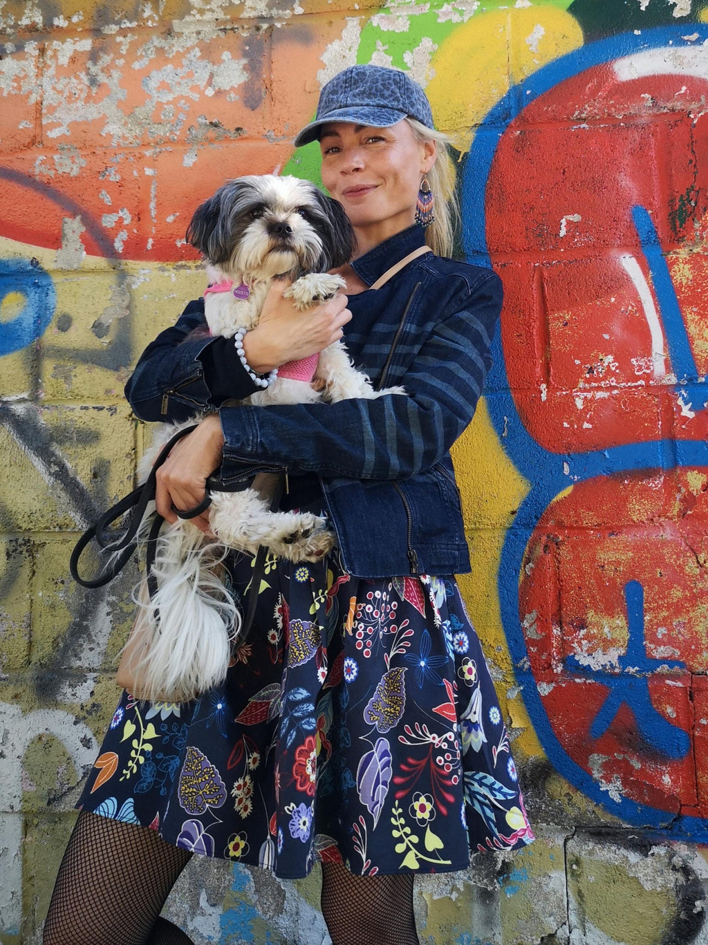 Peut inclure: Une personne tenant un petit chien devant un mur de graffiti coloré. La personne porte une veste en jean, une casquette bleue et une jupe à motifs floraux. Le chien a des poils noirs et blancs et un collier rose.