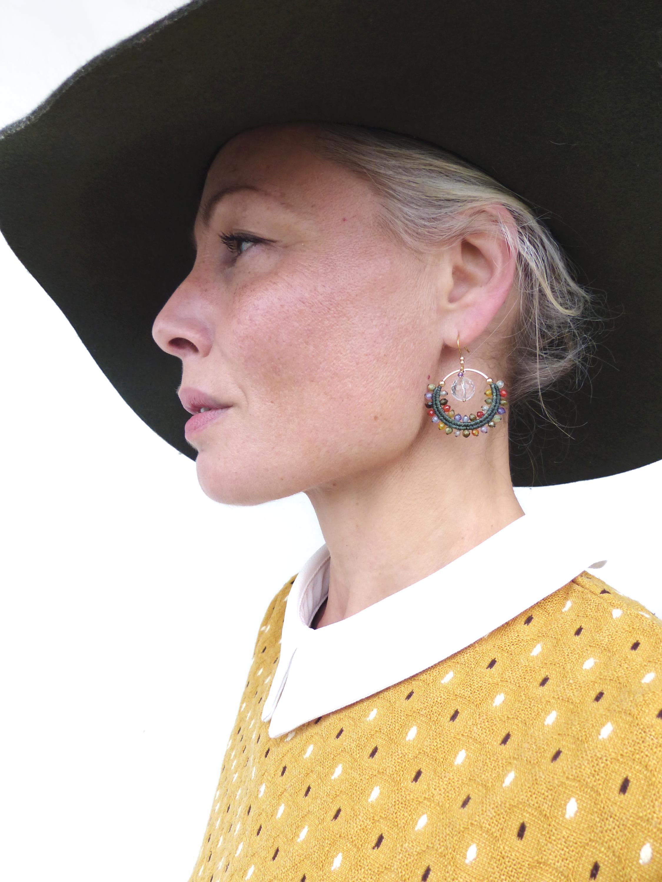 Peut inclure: Une femme porte un chapeau à larges bords vert olive et un pull moutarde à pois blancs. Elle porte des boucles d'oreilles créoles dorées ornées de perles colorées et d'un cristal transparent. Le pull a un col blanc.