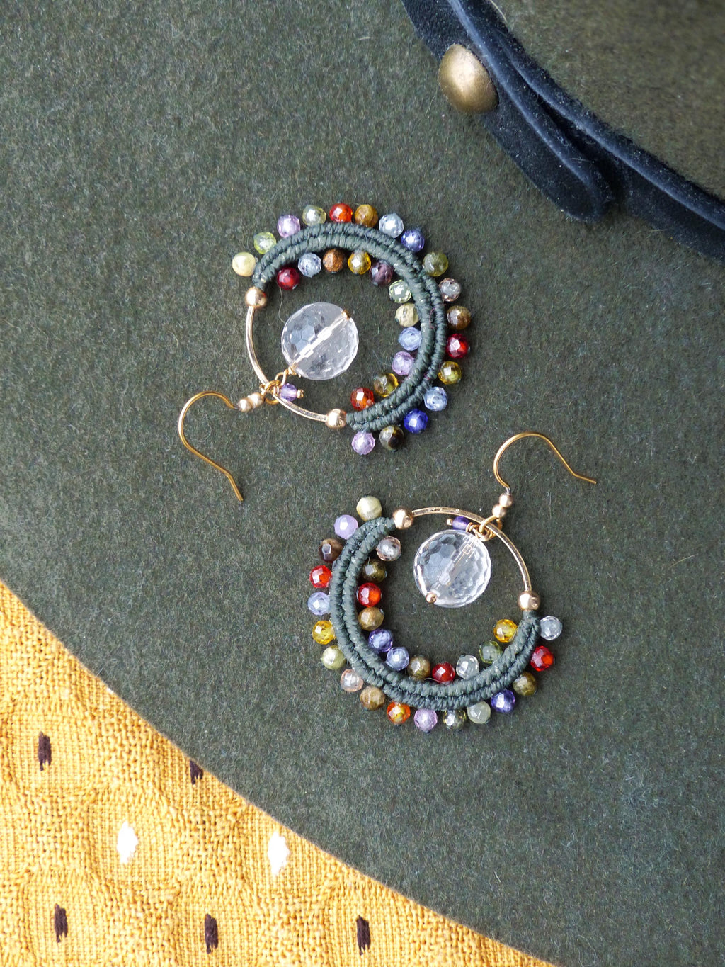 Peut inclure: Une paire de boucles d'oreilles créoles faites à la main. Chaque boucle d'oreille présente un cerceau doré avec un cristal clair et rond et un design macramé de couleur vert foncé. Le macramé est orné de perles colorées de différentes tailles et teintes.