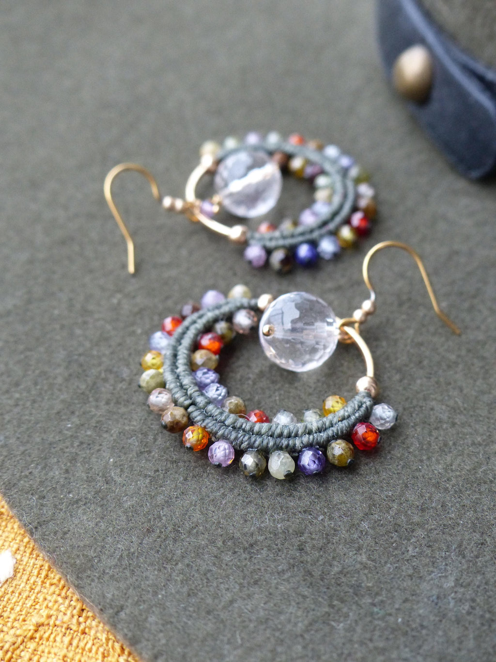 Peut inclure: Une paire de boucles d'oreilles créoles faites à la main. Chaque boucle d'oreille est dotée d'un crochet doré, d'un cercle de style macramé vert et est ornée de petites perles colorées rouges, oranges, violettes et vertes. Une perle transparente et ronde est suspendue au centre.