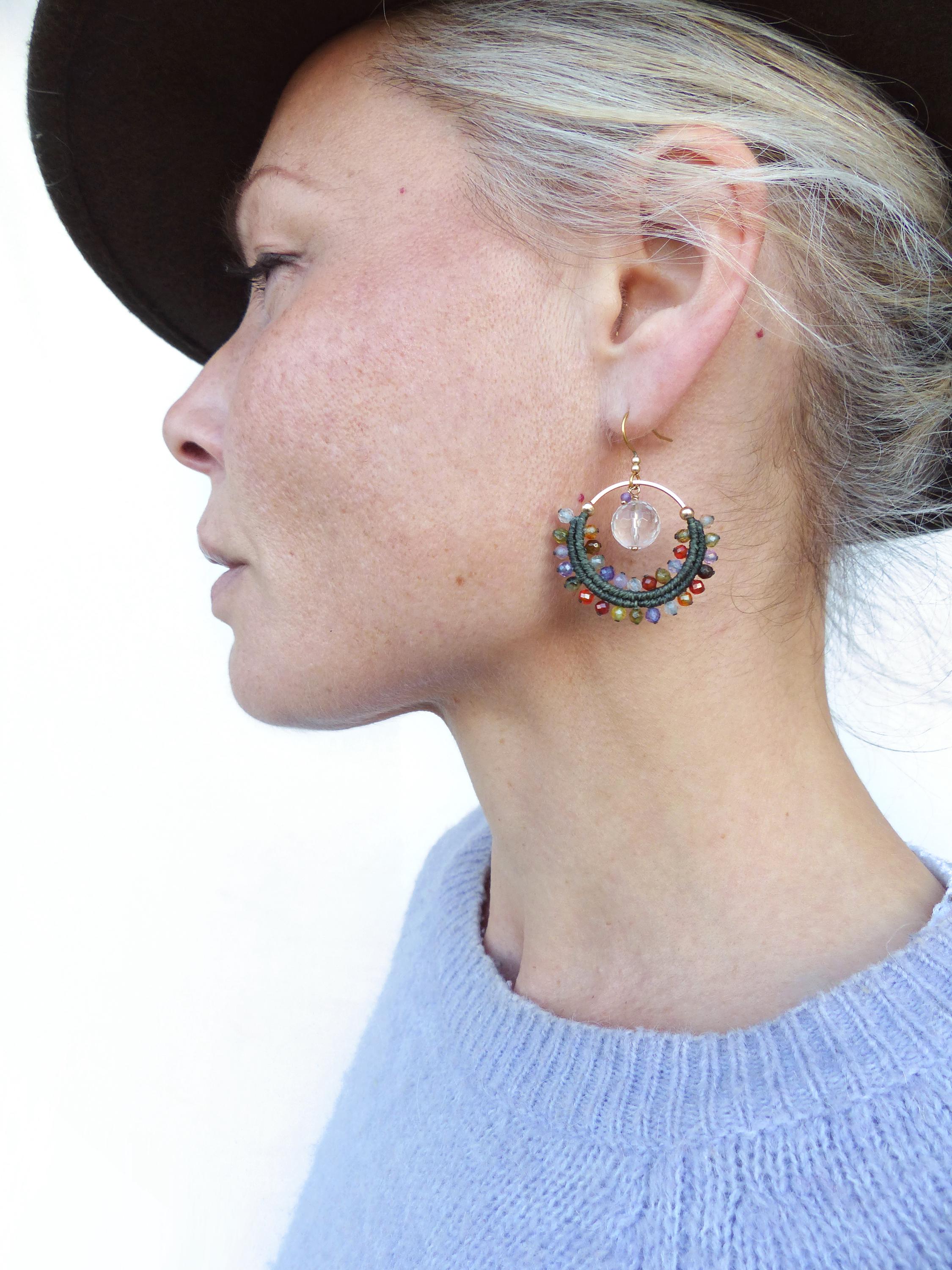 Peut inclure: Boucles d'oreilles créoles dorées avec un design macramé. Les boucles d'oreilles présentent un motif circulaire avec des perles vertes, rouges et transparentes, et une pierre rose centrale. La personne porte un pull bleu et un chapeau marron.