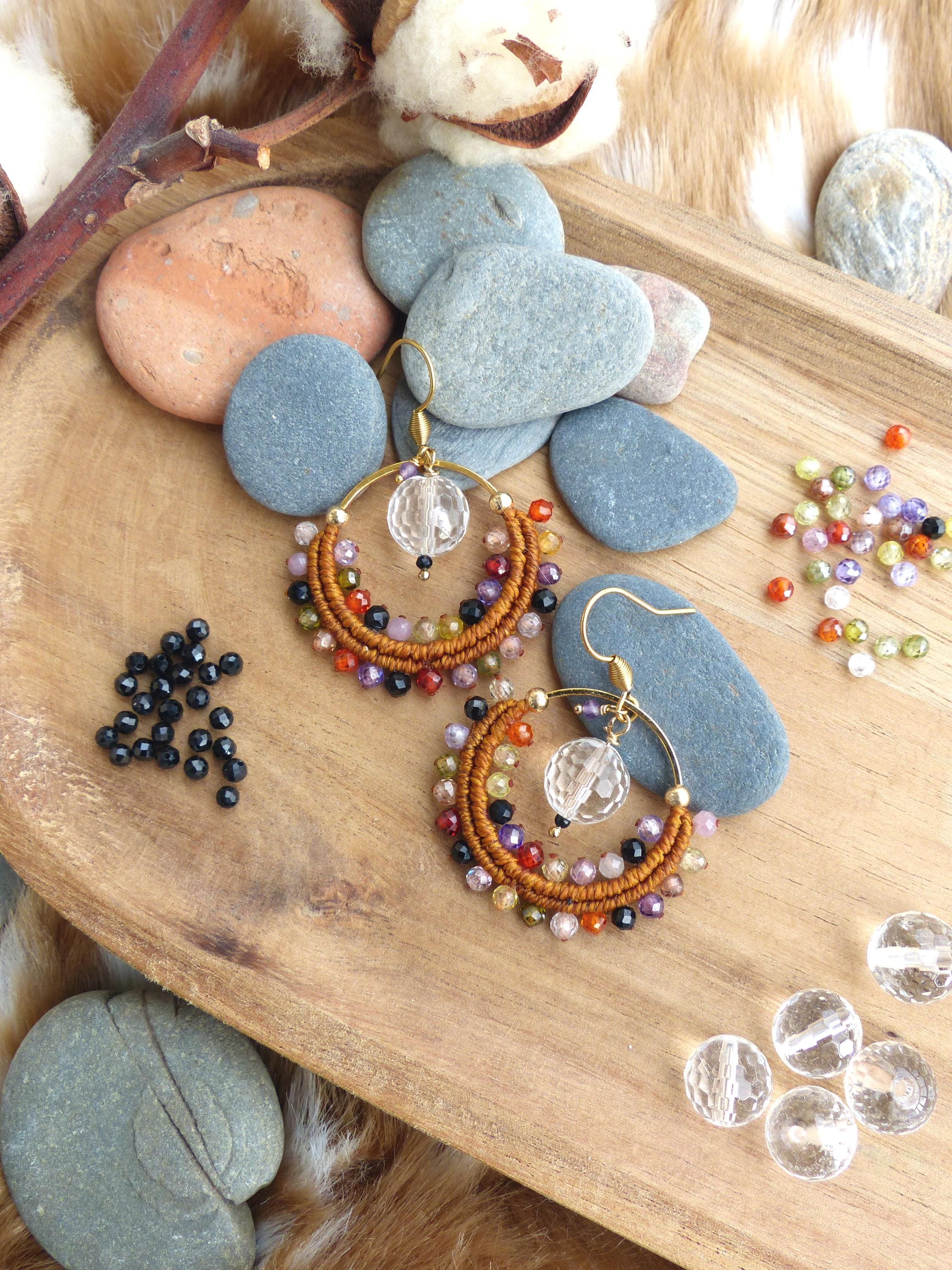 Peut inclure: Paire de boucles d'oreilles créoles faites à la main, avec du métal doré et des détails en macramé. Les boucles d'oreilles présentent un mélange de perles colorées et une perle transparente et facettée au centre. Présentées sur un plateau en bois avec des pierres et des perles décoratives.