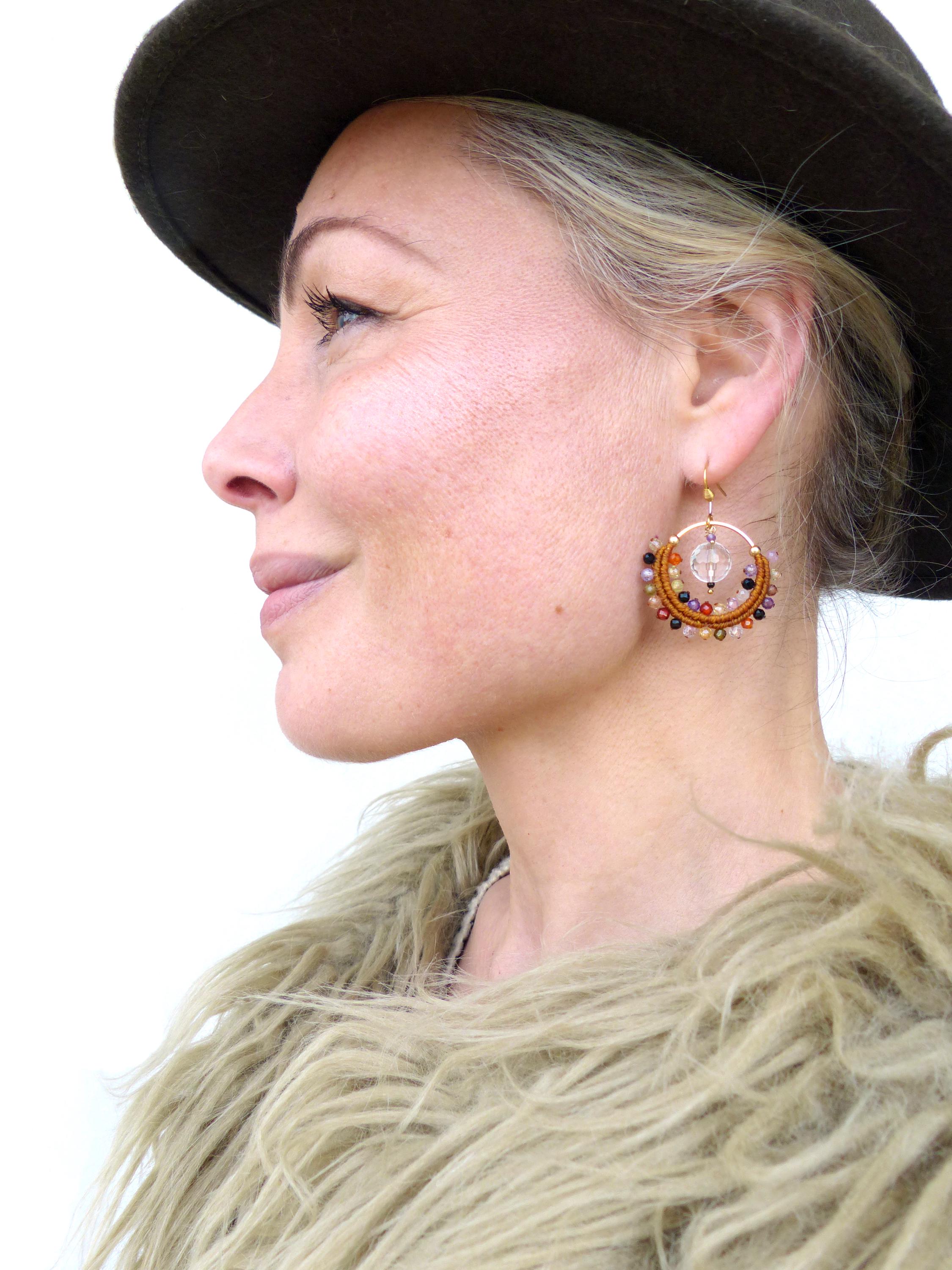 Peut inclure: Une femme porte un chapeau marron et un manteau en fausse fourrure. Elle porte des boucles d'oreilles dorées avec des perles colorées et un cristal clair au centre. Les boucles d'oreilles sont un accessoire unique et artisanal.