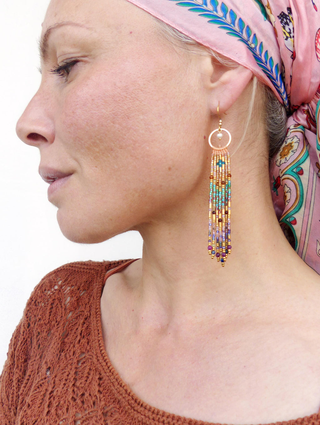 Peut inclure: Gros plan d'une boucle d'oreille dorée avec une frange de perles aux tons violets, turquoise et dorés. La boucle d'oreille est suspendue à un anneau doré. La personne porte un haut rouille et un foulard rose.