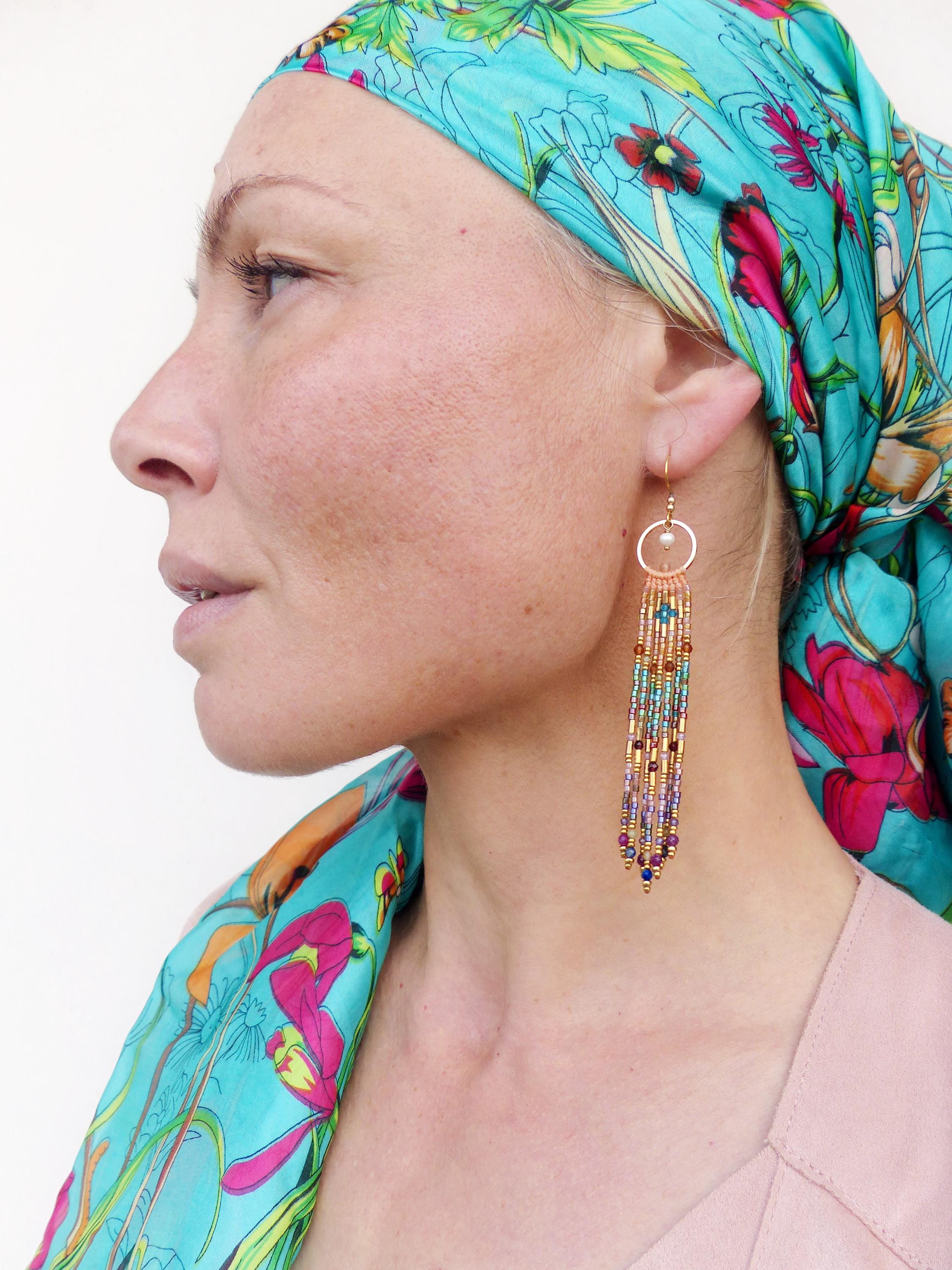 Peut inclure: Boucles d'oreilles pendantes en perles dorées avec un design à franges. Les boucles d'oreilles présentent un petit cercle doré avec une perle et plusieurs brins de perles colorées dans les tons de bleu, violet et or. Les boucles d'oreilles sont longues.
