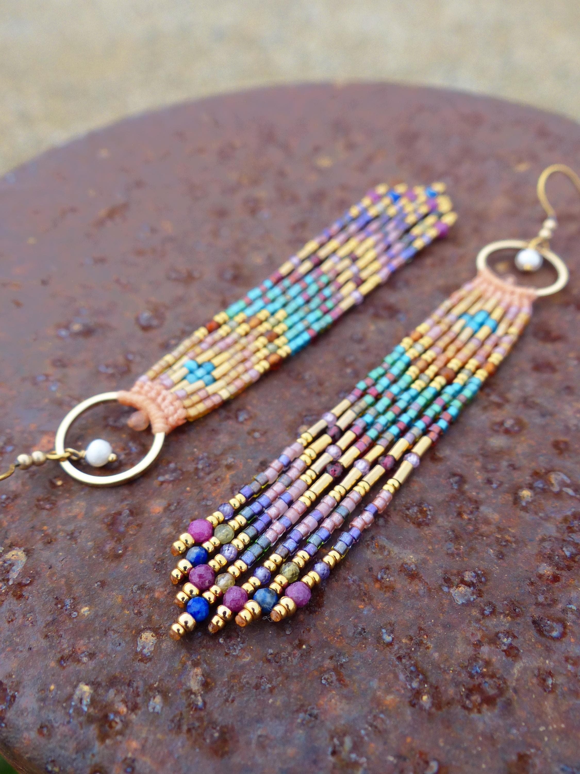 Peut inclure: Boucles d'oreilles faites à la main avec un design à franges. Les boucles d'oreilles présentent un mélange de perles colorées dans les tons de bleu, violet, or et rose. Elles ont des cerceaux dorés et sont présentées sur une surface brun rouillé.