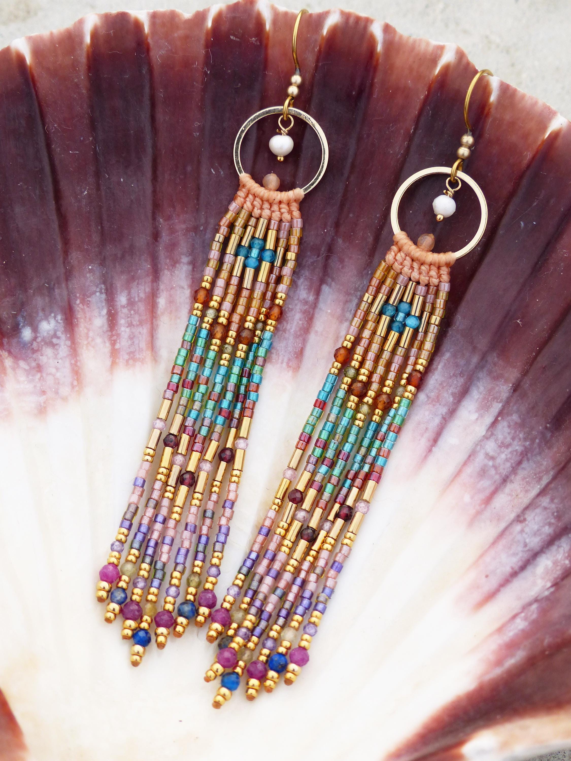 Peut inclure: Une paire de boucles d'oreilles faites à la main avec des perles, avec un anneau doré et un design à franges. Les boucles d'oreilles présentent une variété de perles colorées dans les tons de bleu, vert, violet et or. Les boucles d'oreilles sont présentées sur un coquillage.