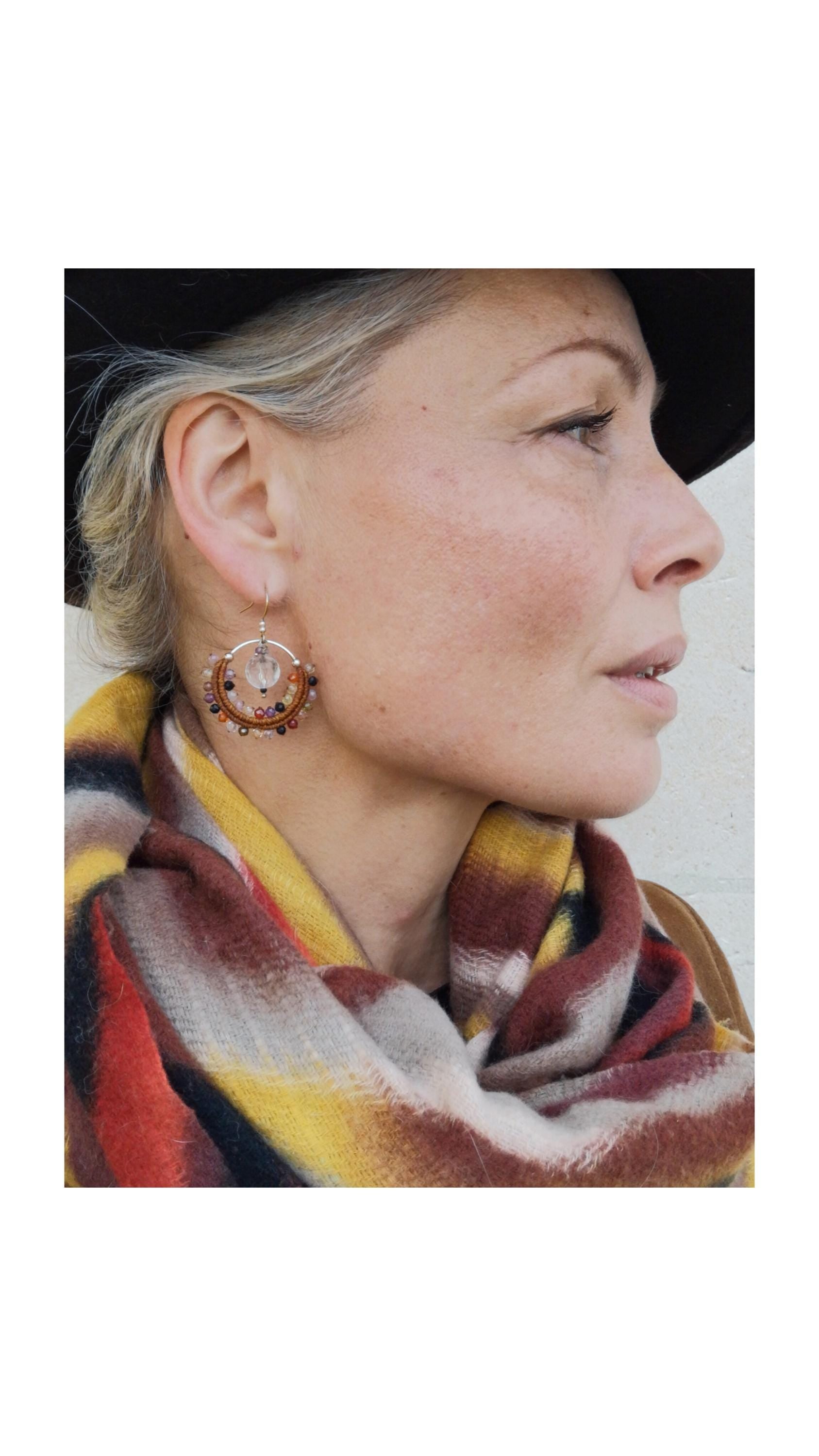 Peut inclure: Une femme porte des boucles d'oreilles créoles ornées de diverses perles et d'une pierre centrale transparente. Elle porte un chapeau noir et une écharpe colorée à rayures rouges, jaunes et marron.