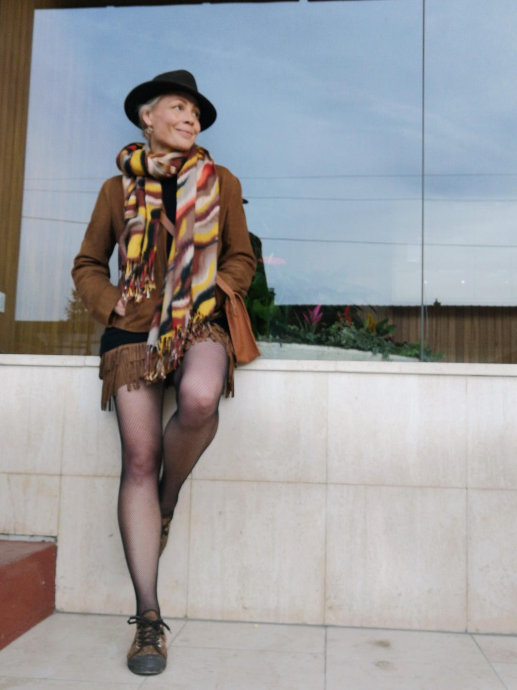 Peut inclure: Une personne porte une veste en daim marron, une écharpe à motifs aux tons jaunes, oranges et bruns, et un chapeau noir. Elle porte également une jupe à franges, des bas noirs et des chaussures marron. Un sac en cuir marron est suspendu à son épaule.
