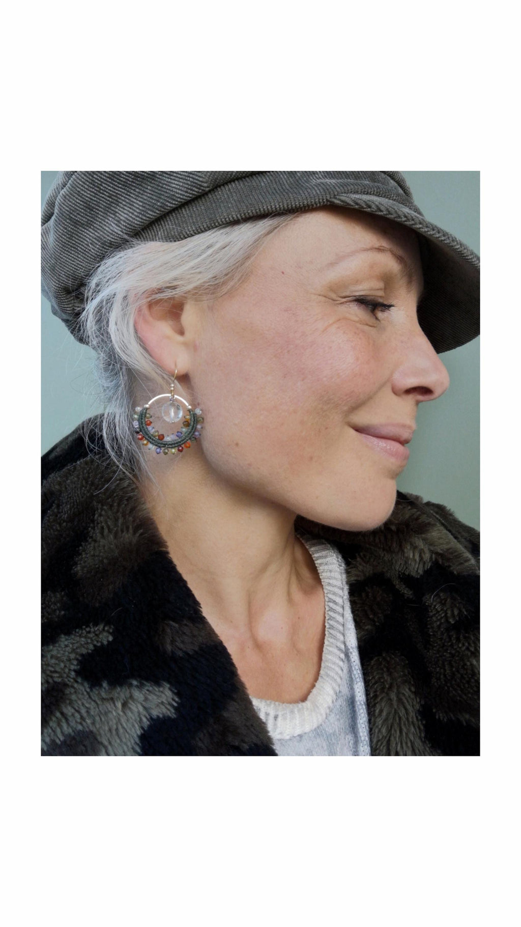 Peut inclure: Boucles d'oreilles créoles argentées avec des perles colorées et une pierre centrale transparente. Les boucles d'oreilles sont portées par une personne portant un chapeau gris et une veste sombre à motifs. Un accessoire unique.