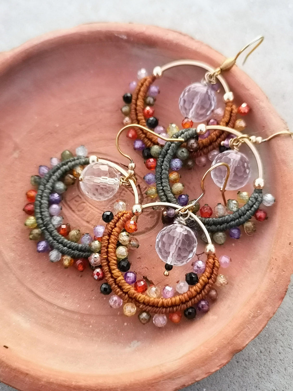 Peut inclure: Gros plan sur trois paires de boucles d'oreilles créoles. Chaque boucle d'oreille présente un cerceau doré avec un motif macramé marron ou gris, orné de perles colorées et d'un pendentif boule de cristal facettée. Les boucles d'oreilles reposent sur une assiette couleur terre cuite.