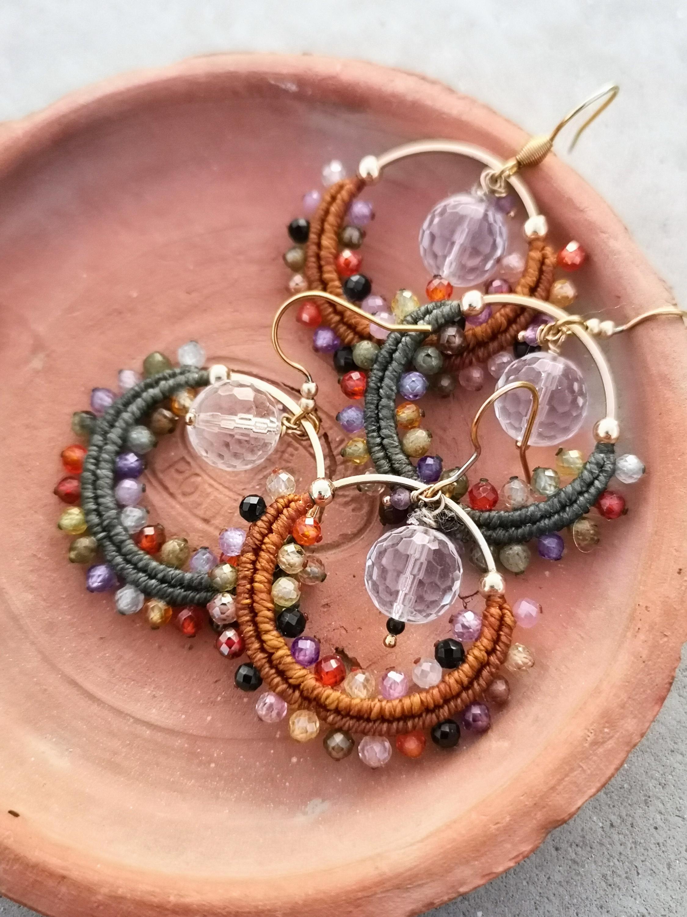 Peut inclure: Gros plan sur trois paires de boucles d'oreilles créoles. Chaque boucle d'oreille présente un cerceau doré avec un motif macramé marron ou gris, orné de perles colorées et d'un pendentif boule de cristal facettée. Les boucles d'oreilles reposent sur une assiette couleur terre cuite.