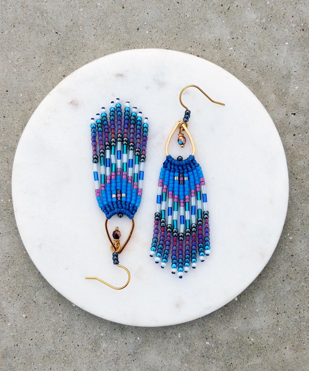 Peut inclure: Une paire de boucles d'oreilles perlées faites à la main. Les boucles d'oreilles présentent un motif de couleurs bleu, violet et blanc. Le design comprend une frange de perles suspendue à un crochet en métal doré. Présentées sur une surface en marbre blanc.
