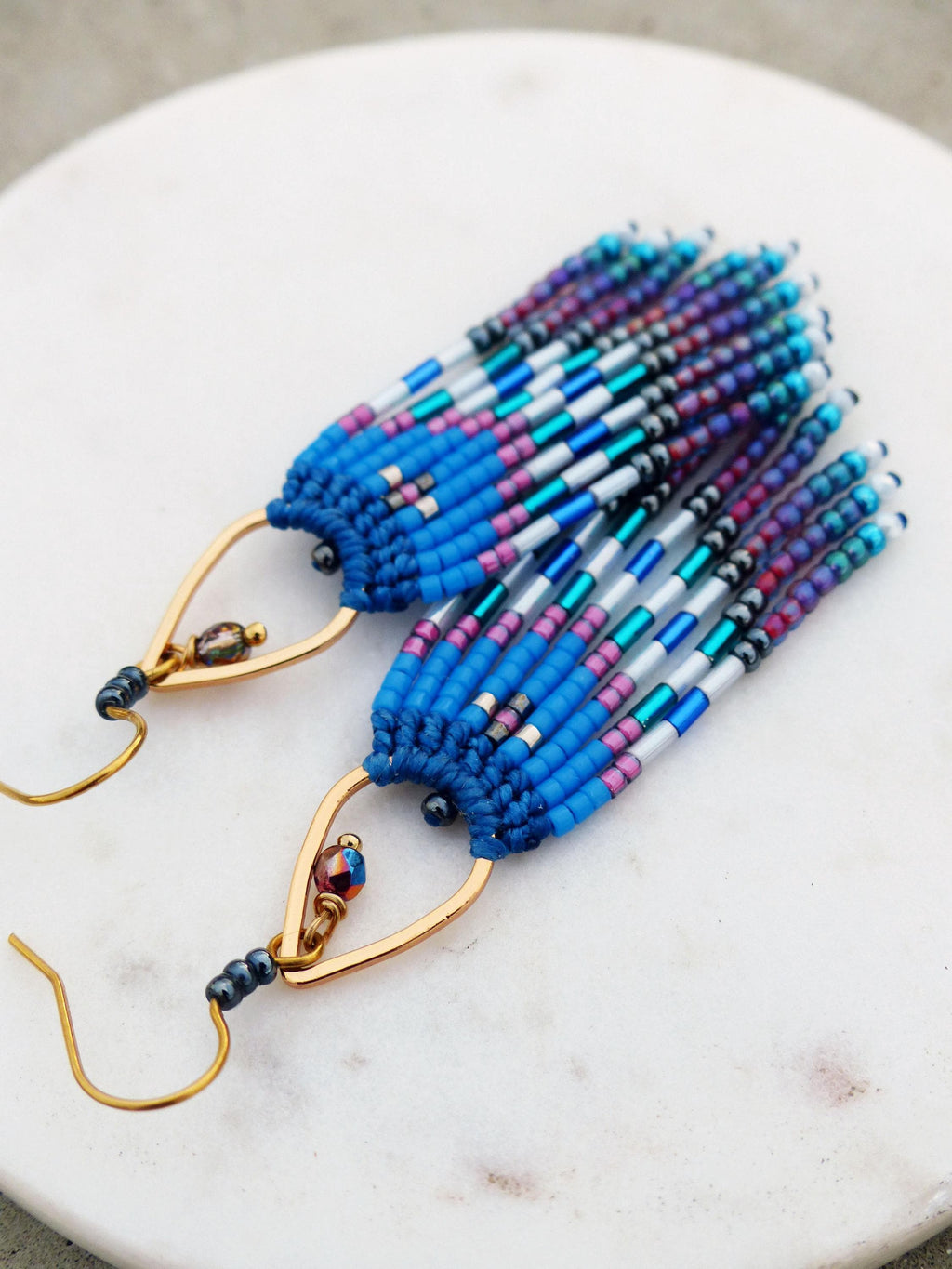 Peut inclure: Boucles d'oreilles faites à la main avec un design à franges. Les boucles d'oreilles présentent un mélange de perles bleues, violettes, argentées et noires, suspendues à des crochets dorés. Elles sont présentées sur une surface en marbre blanc. Mesurent environ 7 cm de long.