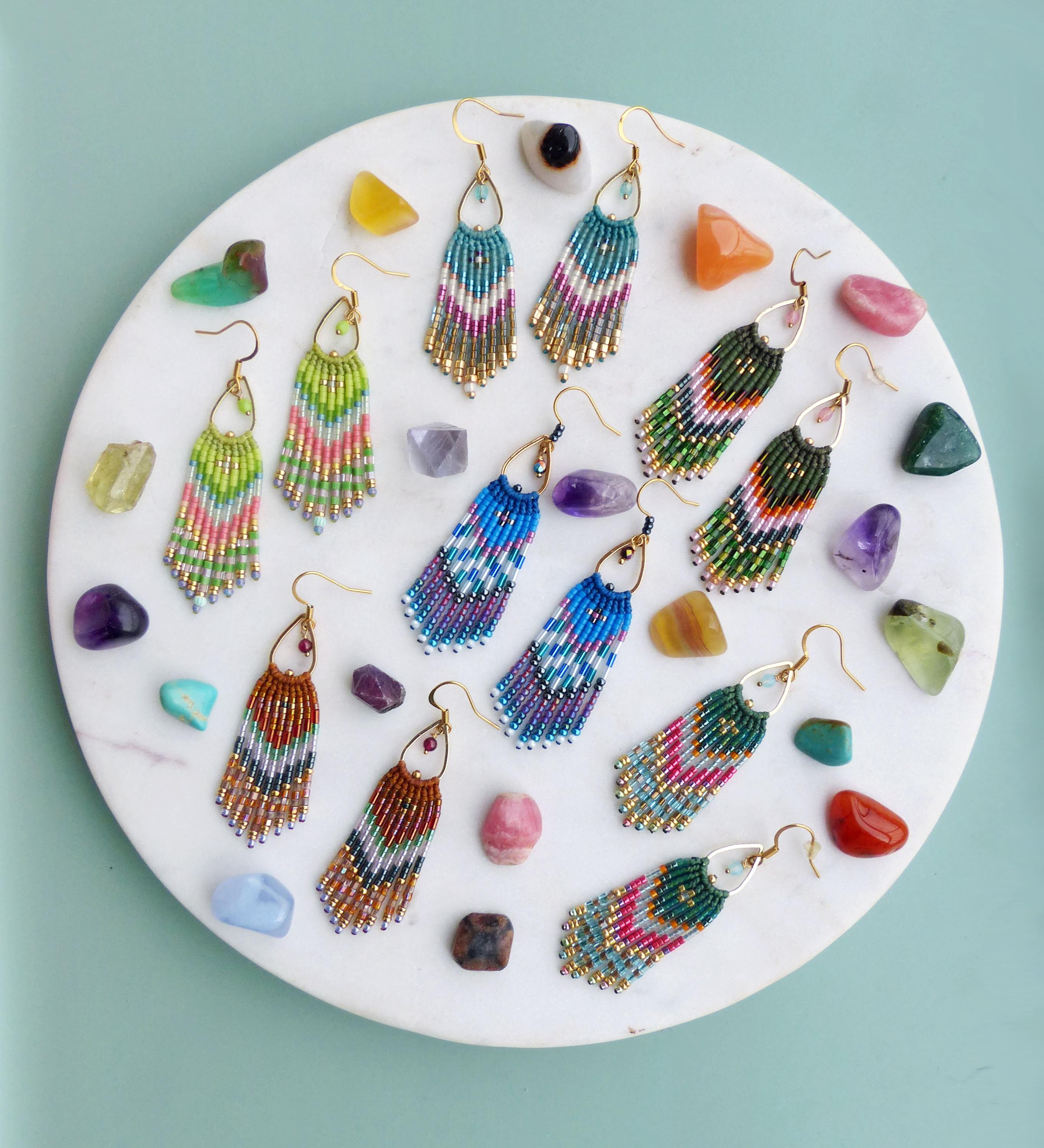 Peut inclure: Une collection de boucles d'oreilles pendantes en perles de différentes couleurs, dont le bleu, le vert, le rose et le marron, avec des accents dorés. Les boucles d'oreilles sont disposées sur une surface en marbre blanc, entourées de pierres précieuses colorées.