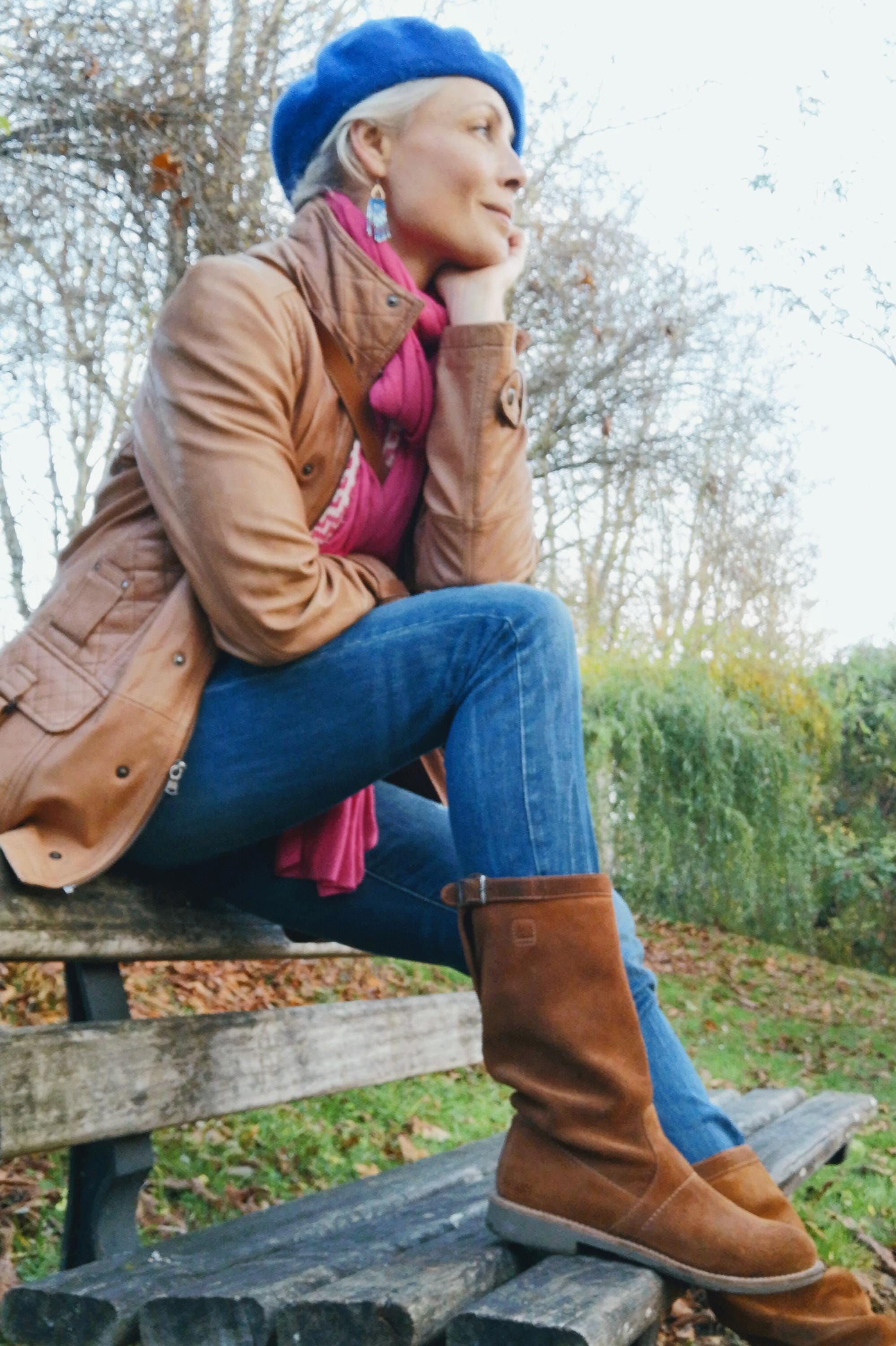 Peut inclure: Une personne portant un béret bleu, une veste en cuir marron, une écharpe rose et un jean bleu. Elle est assise sur un banc en bois, portant des bottes en daim marron. L'arrière-plan montre des arbres et du feuillage.