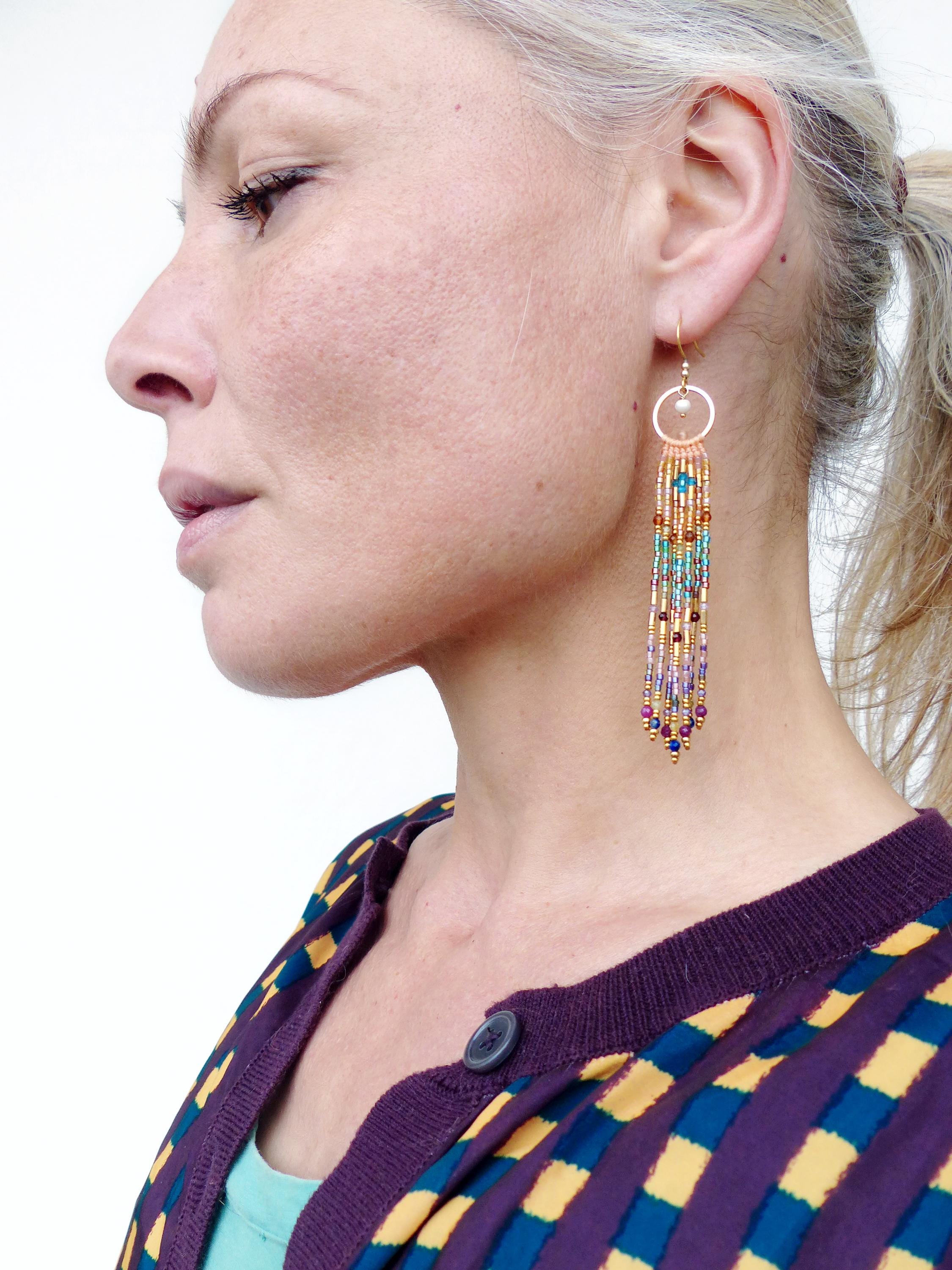 Peut inclure: Boucles d'oreilles en perles dorées avec un design circulaire et des brins de perles colorées pendantes. Les boucles d'oreilles présentent un mélange de perles turquoise, dorées, violettes et rouges, créant un accessoire vibrant. La personne porte un cardigan violet.