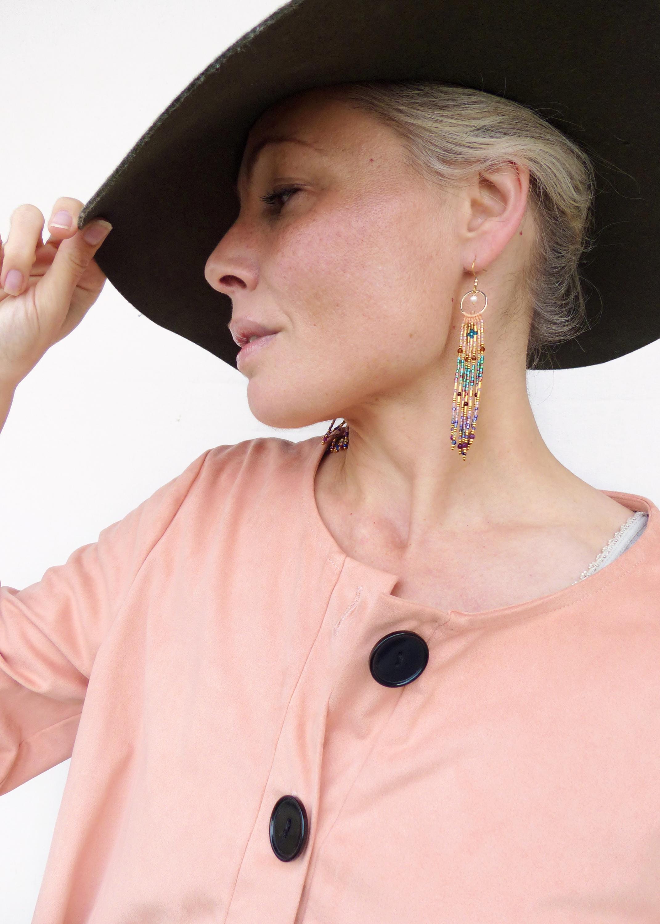 Peut inclure: Une femme porte de longues boucles d'oreilles perlées avec des crochets dorés et une veste pêche. Les boucles d'oreilles présentent des perles turquoise, violettes et dorées. Un chapeau vert foncé est visible en arrière-plan.