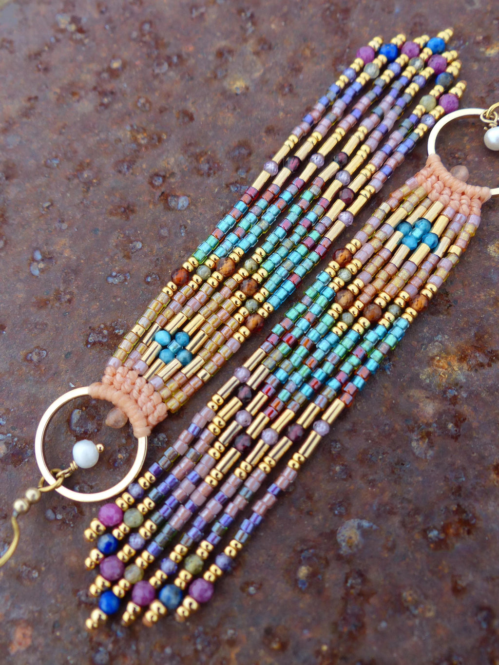 Peut inclure: Une paire de boucles d'oreilles perlées au design géométrique coloré. Les boucles d'oreilles présentent un mélange de perles dorées, bleues, violettes et vertes, avec un anneau doré et une petite perle blanche. Elles sont posées sur une surface brune texturée.