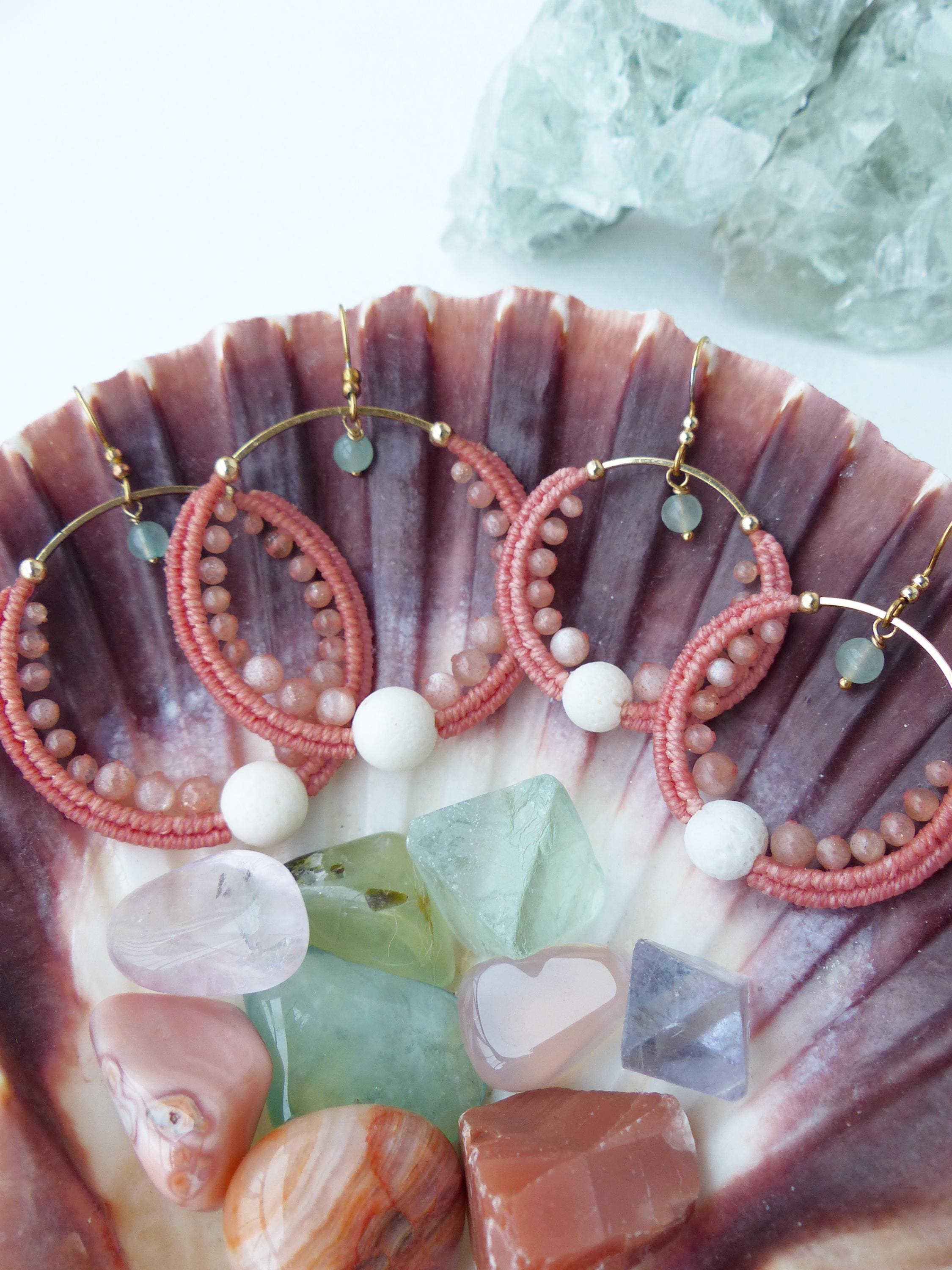 Peut inclure: Quatre paires de boucles d'oreilles créoles avec du macramé rose et de petites perles roses. Chaque boucle d'oreille a un cerceau doré et une perle blanche en bas. Les boucles d'oreilles sont présentées dans un coquillage avec des pierres précieuses colorées.