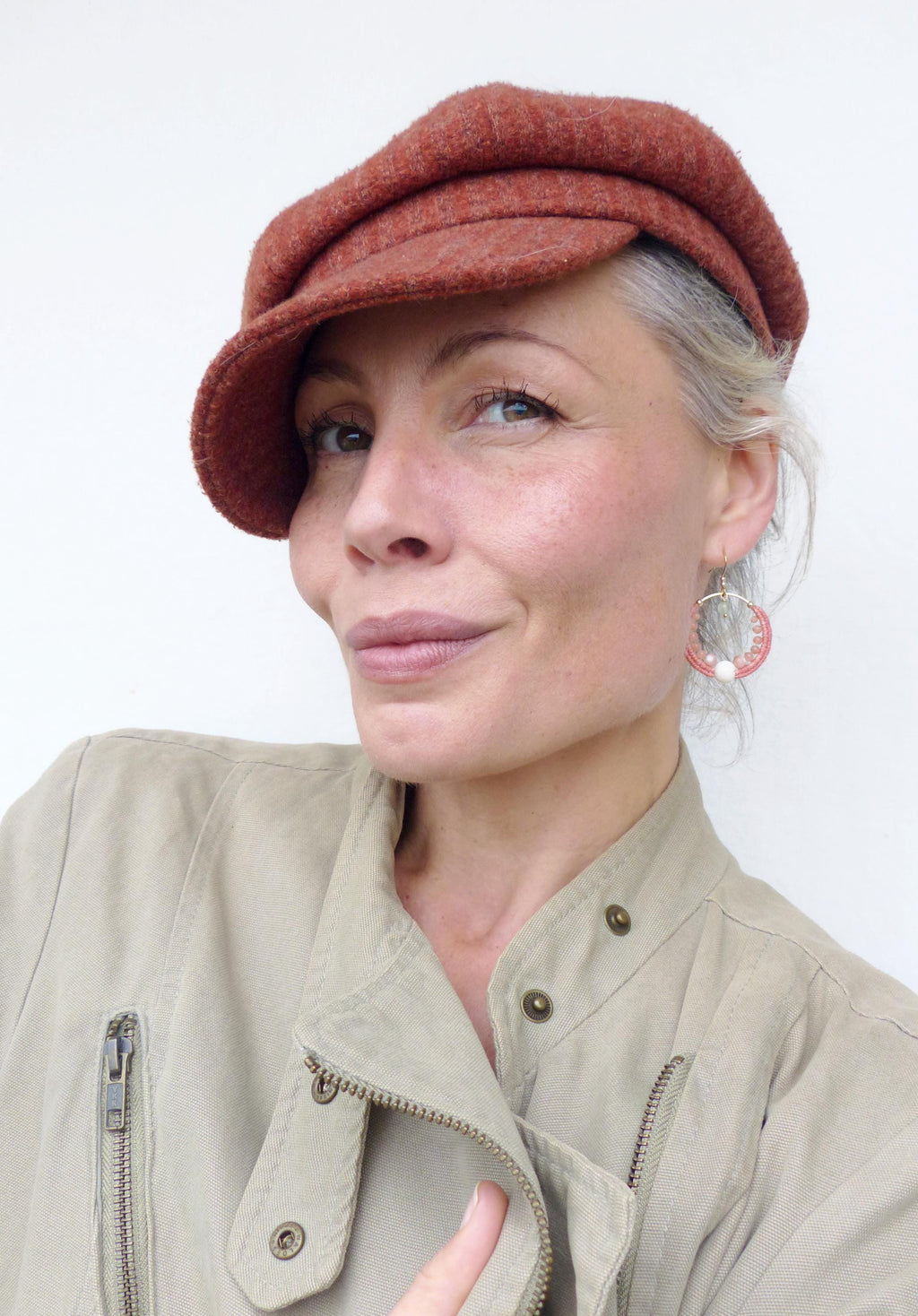 Peut inclure: Une femme porte une casquette de style gavroche rouille et des boucles d'oreilles créoles dorées avec des accents roses et blancs. Elle porte également une veste beige avec une fermeture éclair. Le fond est un mur blanc uni.