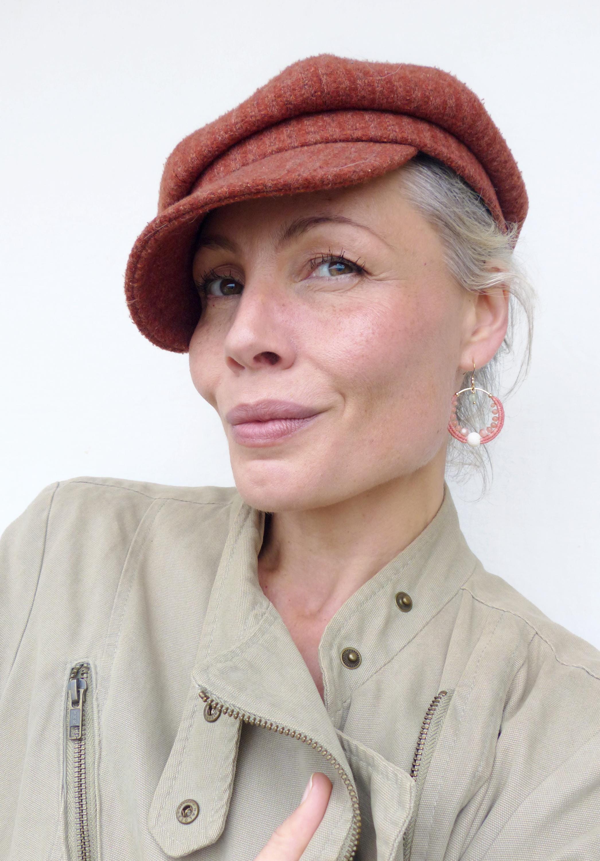 Peut inclure: Une femme porte une casquette de style gavroche rouille et des boucles d'oreilles créoles dorées avec des accents roses et blancs. Elle porte également une veste beige avec une fermeture éclair. Le fond est un mur blanc uni.