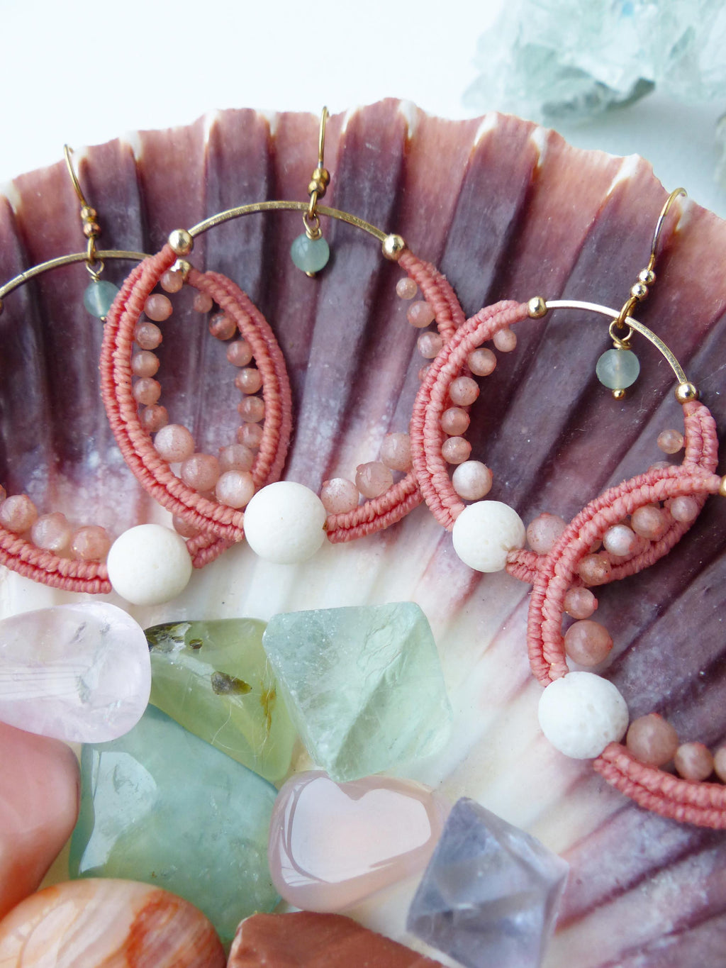 Peut inclure: Gros plan sur trois paires de boucles d'oreilles faites à la main. Chaque boucle d'oreille présente un cerceau doré avec un motif tissé rose, de petites perles roses et une perle blanche. Les boucles d'oreilles sont présentées sur un coquillage, entourées de pierres précieuses colorées.