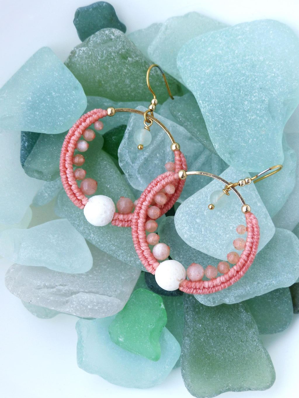 Peut inclure: Une paire de boucles d'oreilles créoles faites à la main avec un motif tissé rose corail. Les boucles d'oreilles comportent de petites perles roses et une grosse perle blanche. Les crochets d'oreilles sont dorés. Les boucles d'oreilles sont présentées sur un lit de verre de mer.
