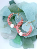 Peut inclure: Une paire de boucles d'oreilles créoles faites à la main avec un motif tissé rose corail. Les boucles d'oreilles comportent de petites perles roses et une grosse perle blanche. Les crochets d'oreilles sont dorés. Les boucles d'oreilles sont présentées sur un lit de verre de mer.