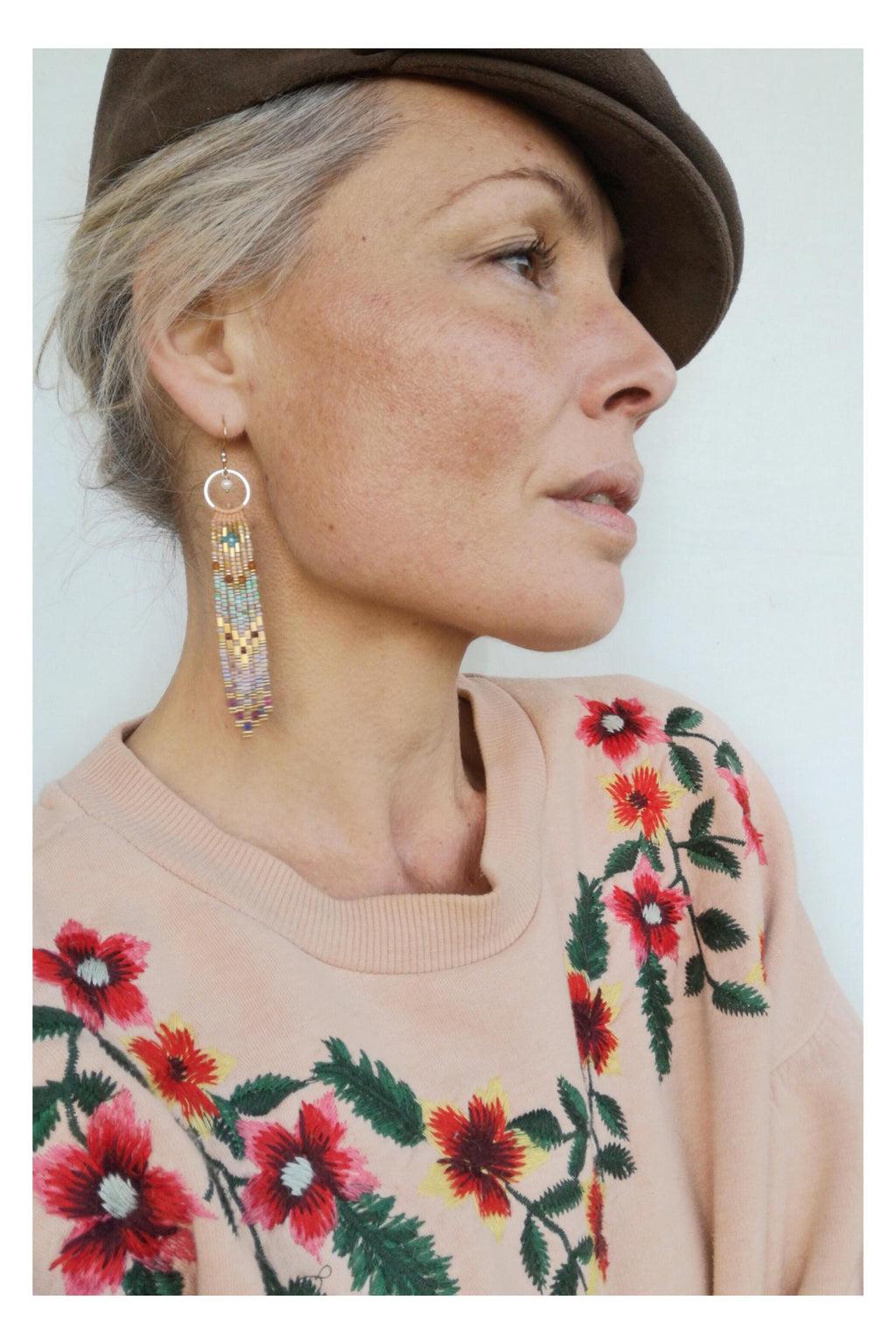 Peut inclure: Une femme porte de longues boucles d'oreilles en perles avec des perles dorées, turquoise et lavande. Elle porte un sweat-shirt rose clair avec des broderies florales rouges et jaunes et une casquette marron.