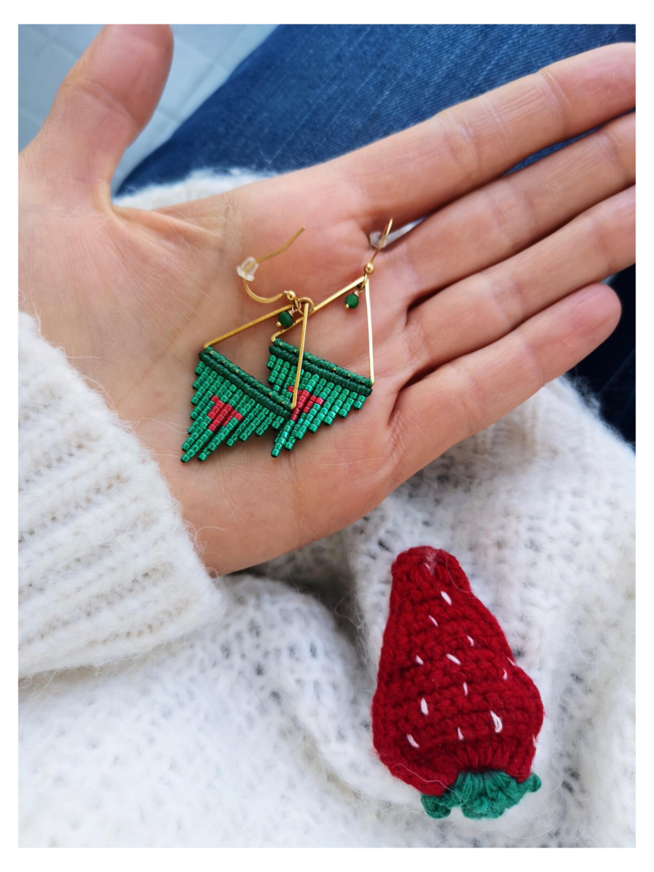 Peut inclure: Boucles d'oreilles triangulaires dorées avec des détails perlés verts et rouges. Une épingle fraise rouge crochetée avec des détails de graines blanches repose sur un pull blanc. Les boucles d'oreilles sont tenues dans une main.