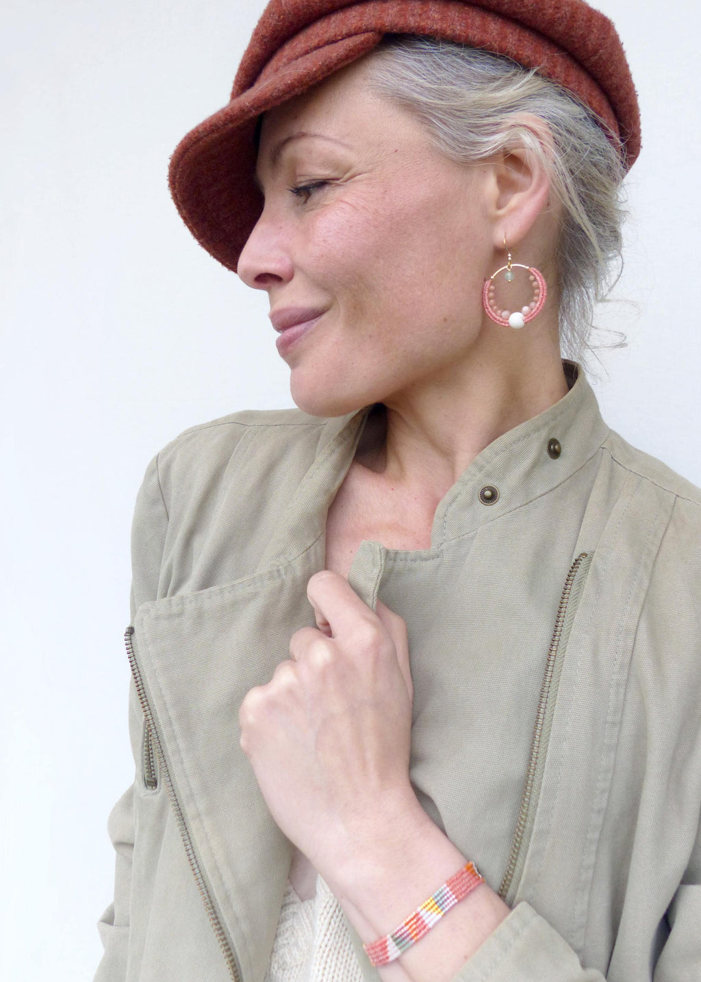 Peut inclure: Une femme porte un chapeau rouille, des boucles d'oreilles créoles dorées avec des perles roses et une perle blanche, une veste beige et un bracelet coloré. Le bracelet a un motif tissé rose, jaune, bleu et blanc.