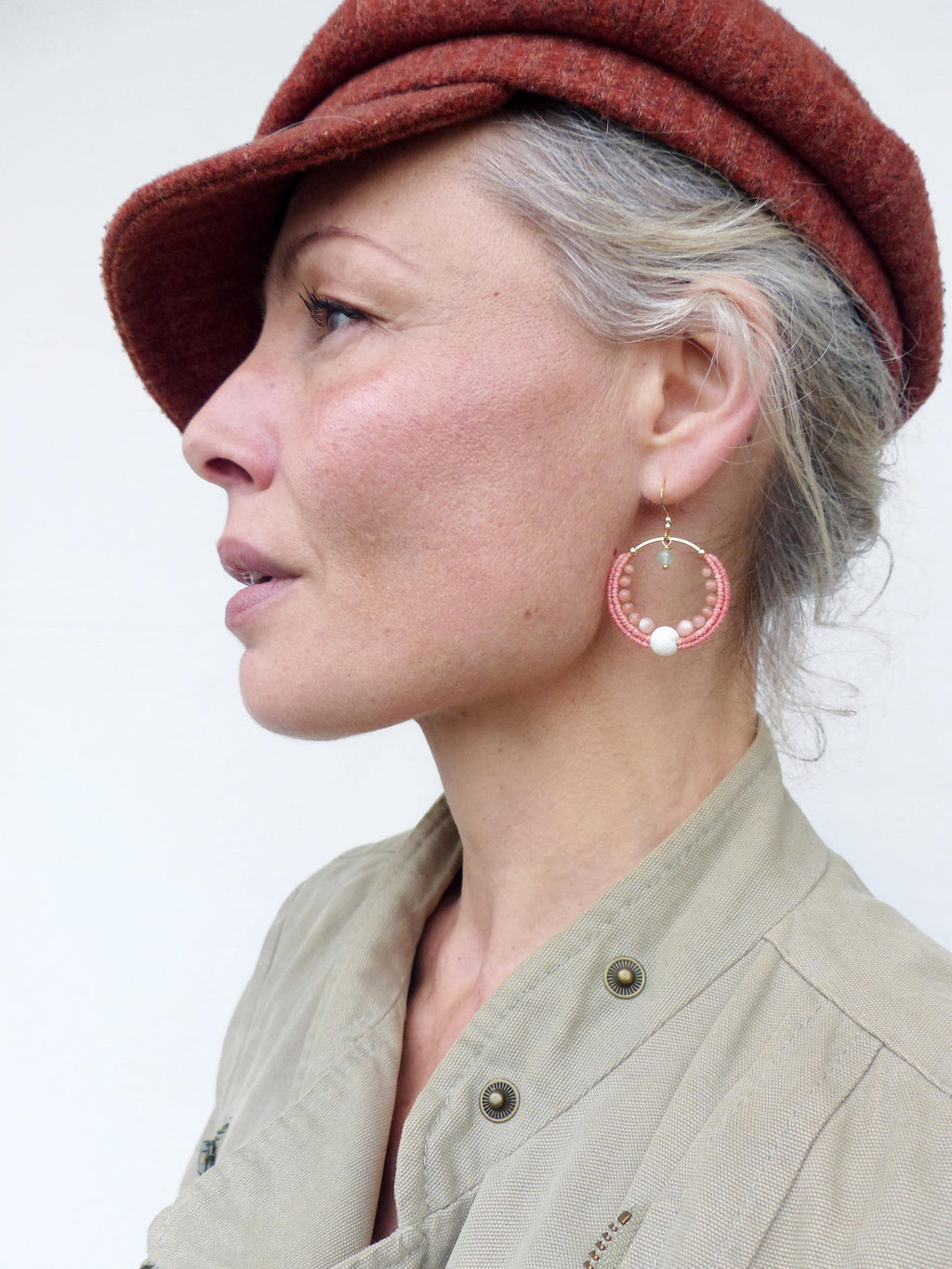 Peut inclure: Une femme porte une casquette de style gavroche rouille et une veste vert clair. L'attention se porte sur une paire de boucles d'oreilles créées à la main. Les boucles d'oreilles présentent un motif rose tissé avec des perles blanches et vert clair, suspendues à des crochets dorés.