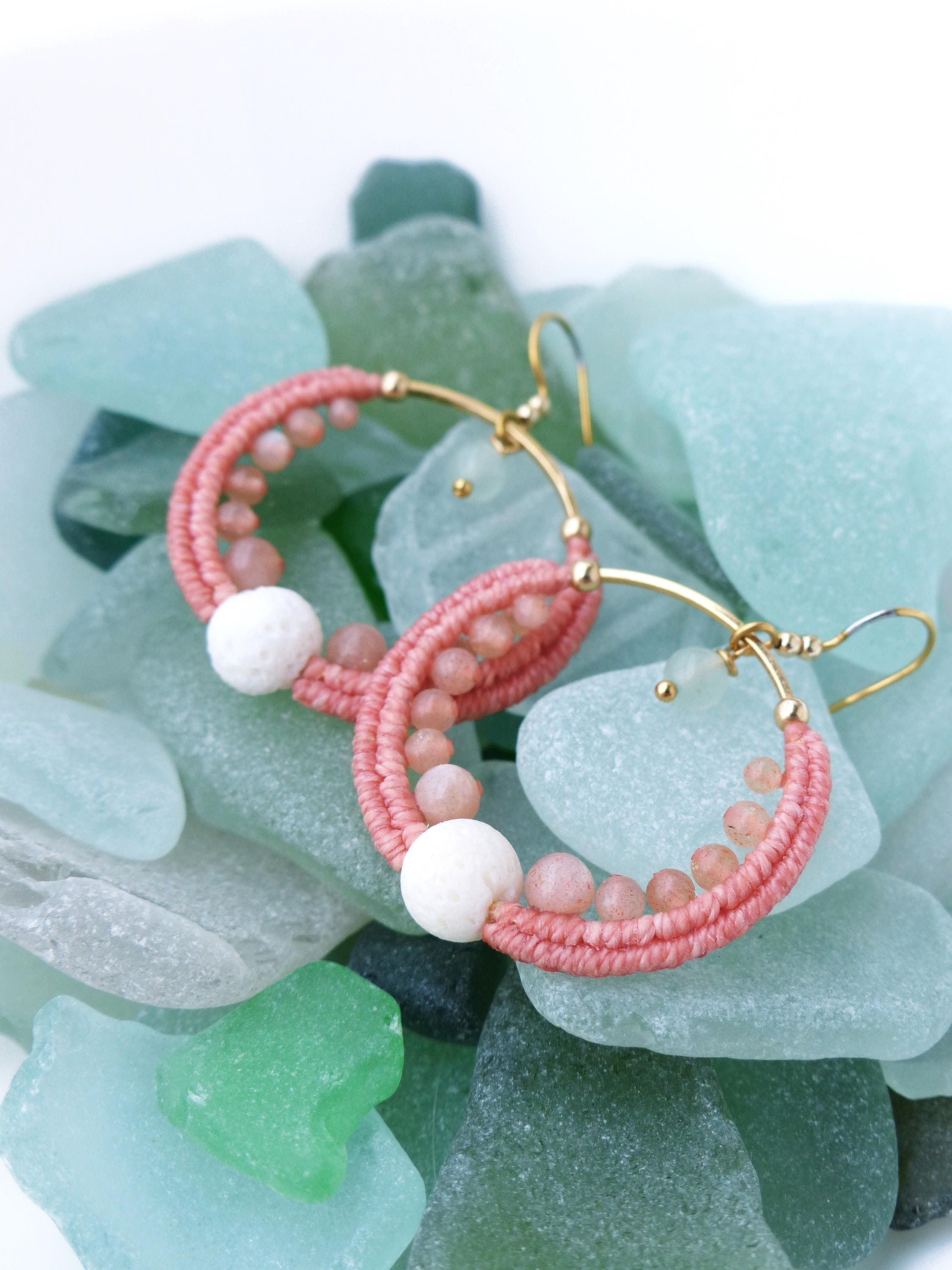 Peut inclure: Une paire de boucles d'oreilles créoles roses avec des crochets et des détails dorés. Les créoles sont enveloppées de fil rose et ornées de petites perles roses et d'accents sphériques blancs. Les boucles d'oreilles reposent sur un lit de verre de mer.