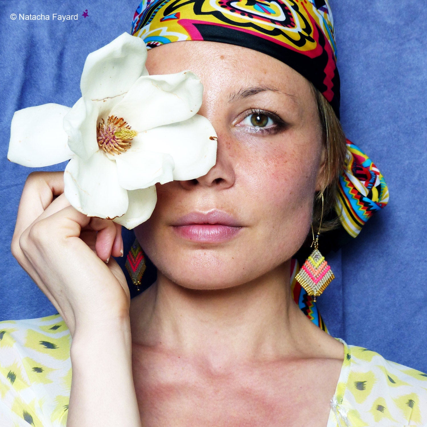 Peut inclure: Une femme portant un foulard coloré et des boucles d'oreilles pose avec une fleur de magnolia blanche qui lui couvre un œil. 