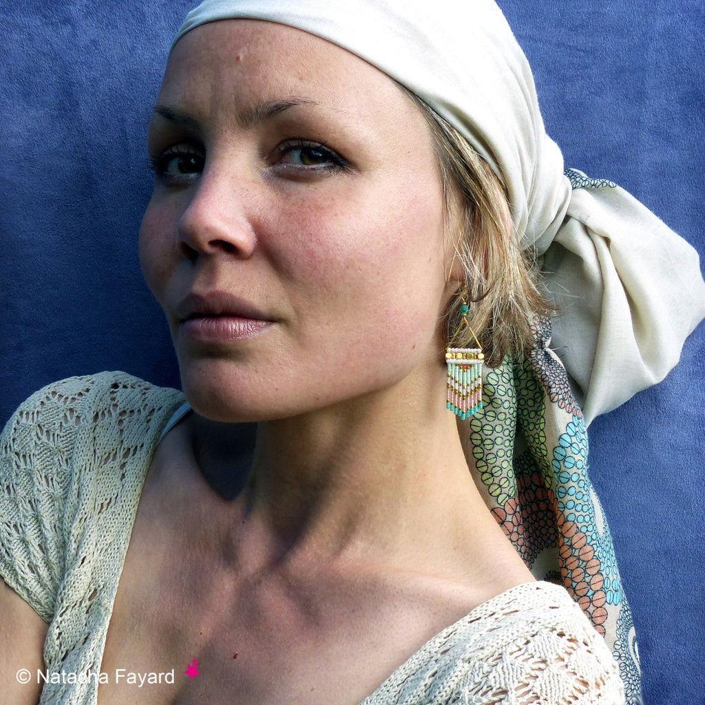 Peut inclure: Une femme portant un foulard blanc et un foulard à motifs vert clair et blanc noué autour du cou. Elle porte un haut en crochet blanc et des boucles d'oreilles en perles dorées et turquoise.