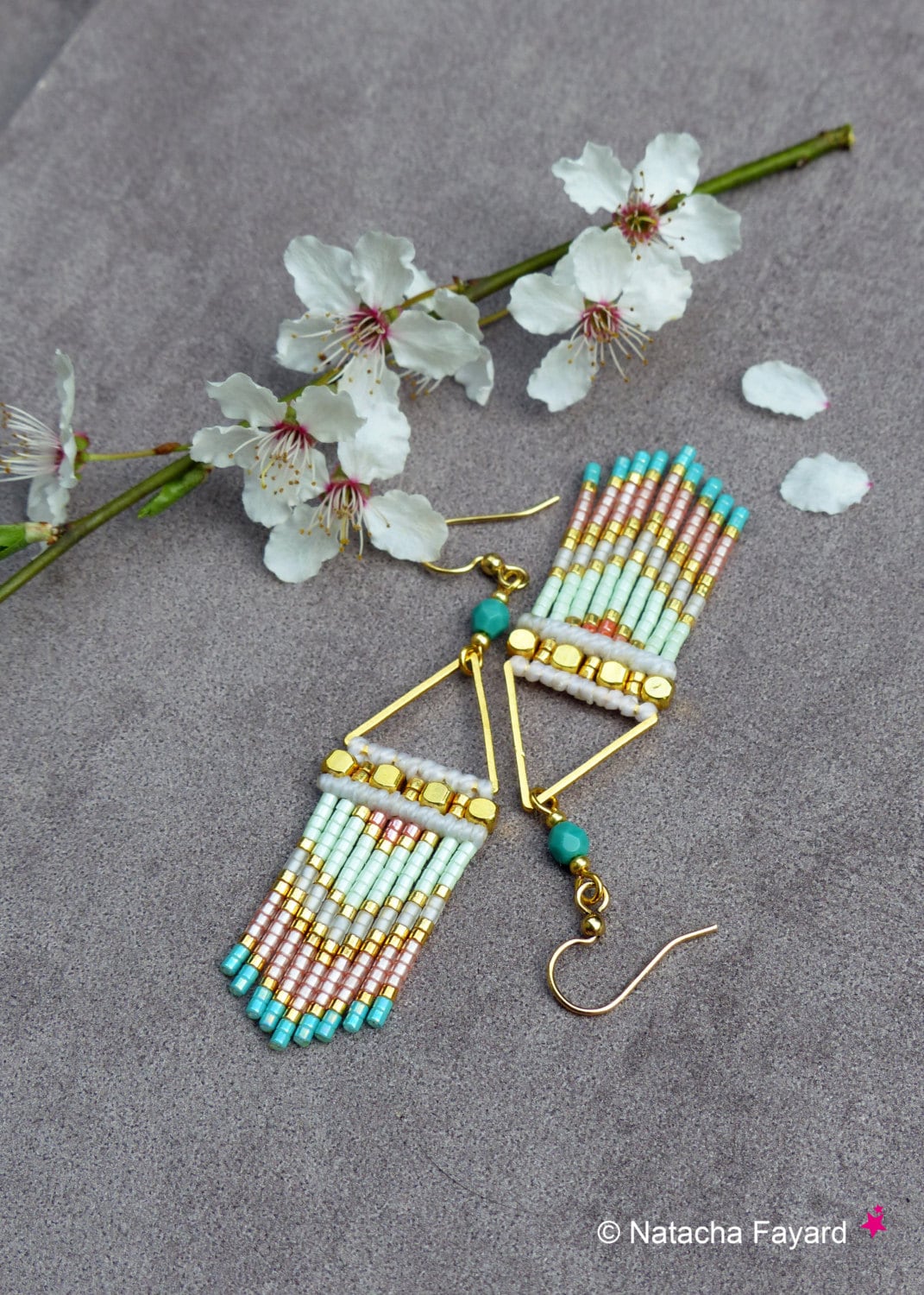 Peut inclure: Une paire de boucles d'oreilles triangulaires dorées avec une frange de perles dans des tons turquoise, blanc, rose et or. La frange est faite de petites perles de rocaille et pend du triangle.