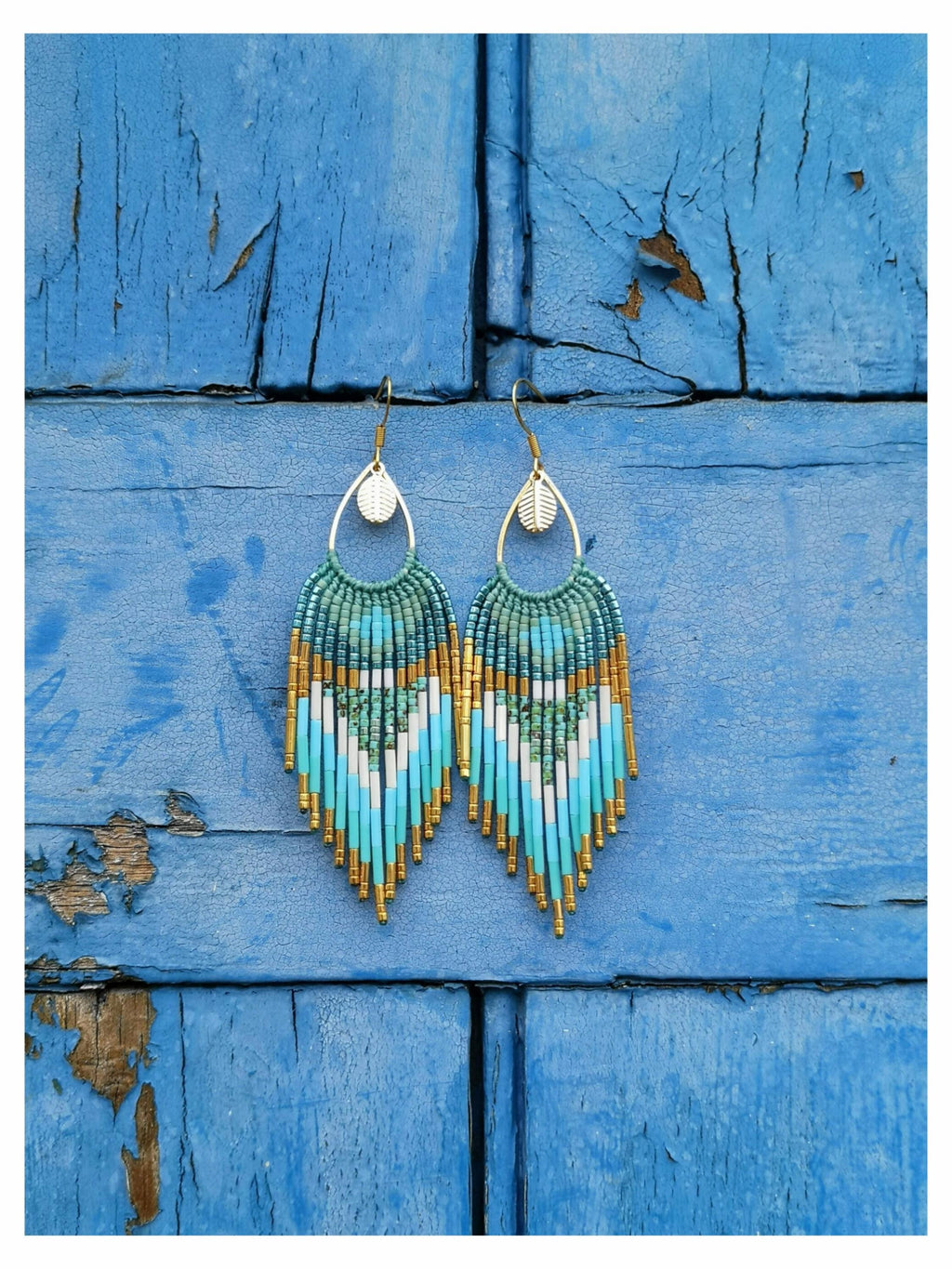 Peut inclure: Paire de boucles d'oreilles faites à la main avec un motif de perles turquoise, blanches et dorées. Les boucles d'oreilles ont une frange de perles dorées en bas et sont suspendues à des crochets dorés avec un charme en forme de feuille. Fond en bois bleu vieilli.