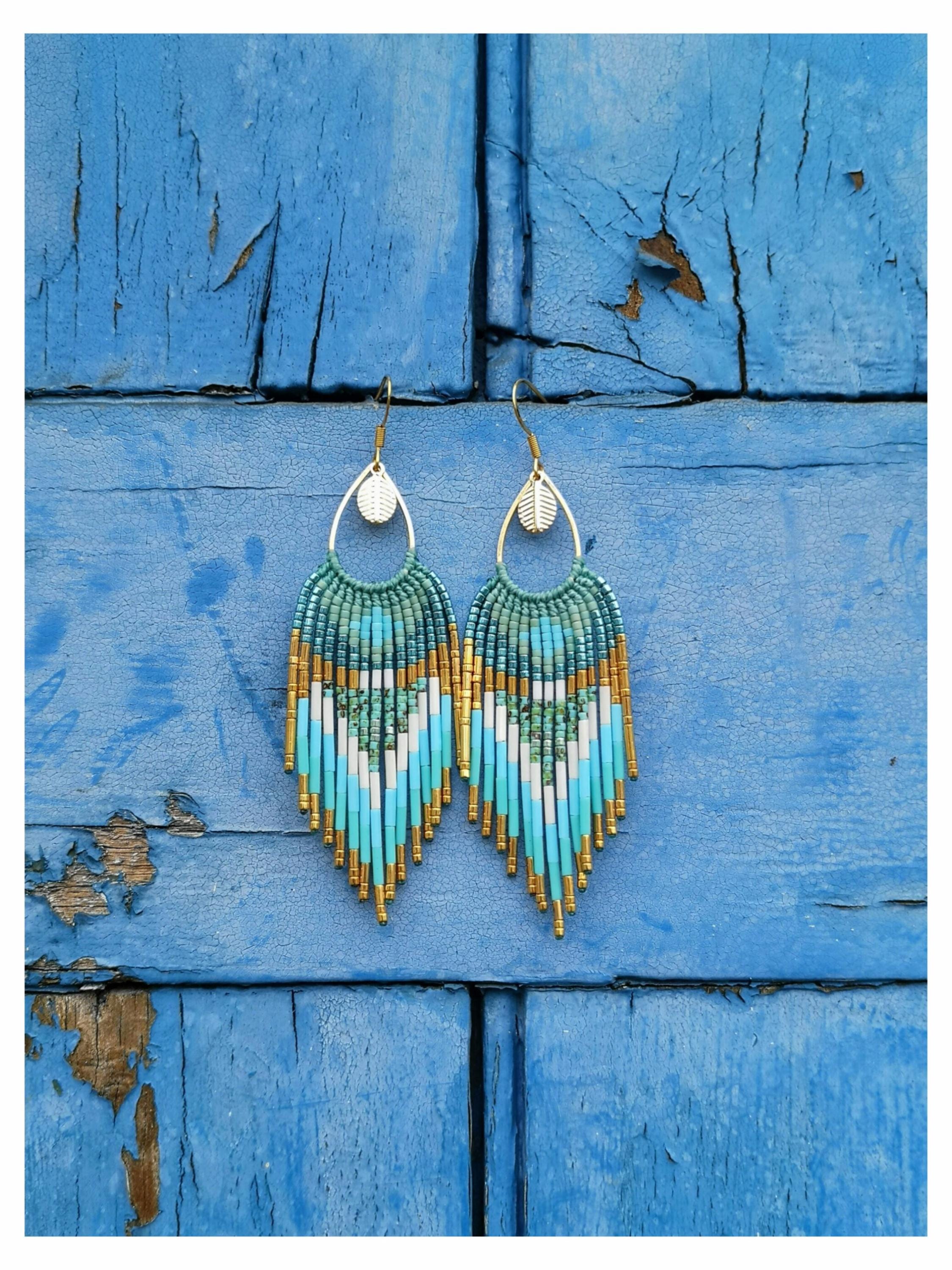 Peut inclure: Paire de boucles d'oreilles faites à la main avec un motif de perles turquoise, blanches et dorées. Les boucles d'oreilles ont une frange de perles dorées en bas et sont suspendues à des crochets dorés avec un charme en forme de feuille. Fond en bois bleu vieilli.